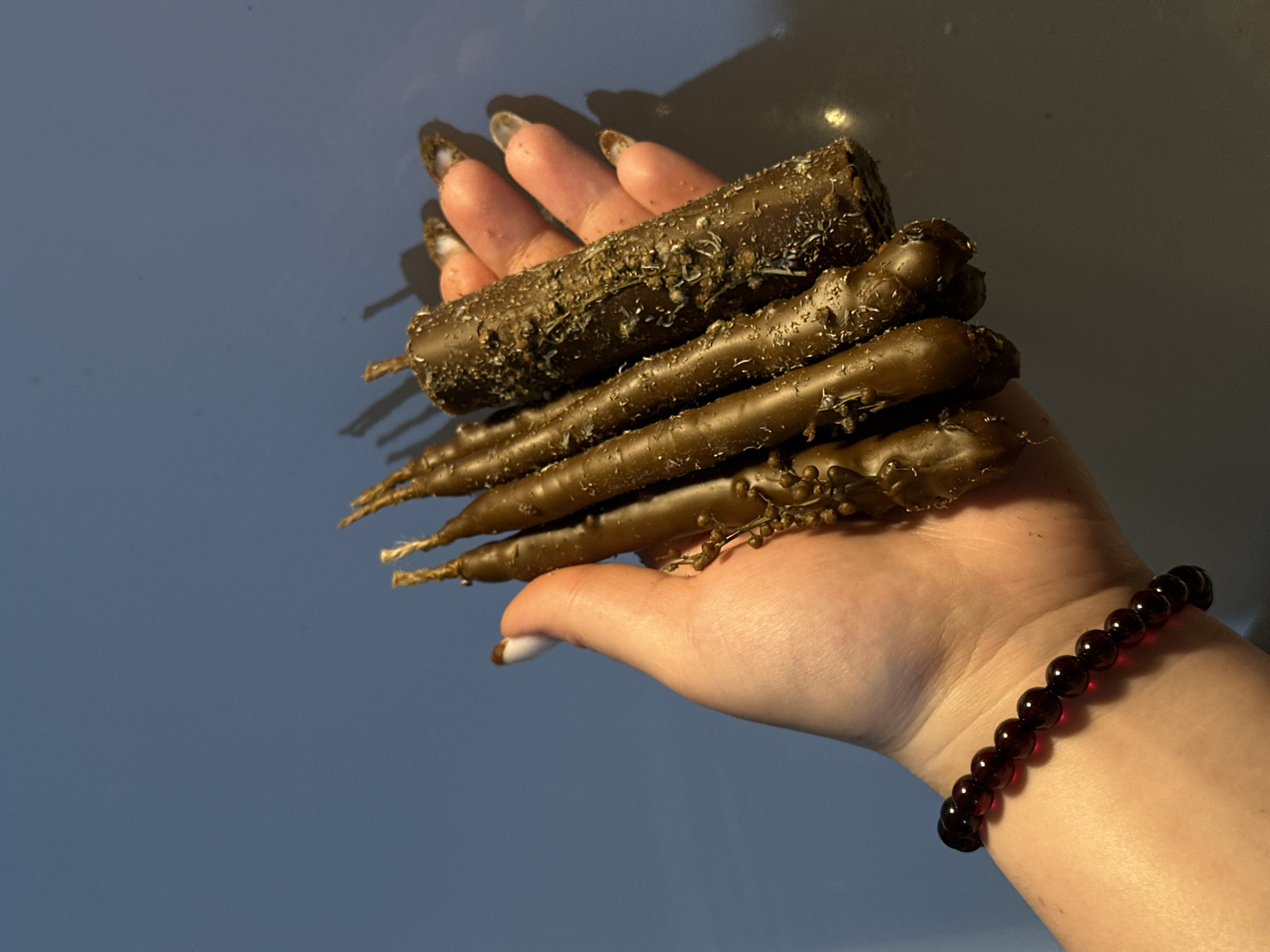 A person's hand holds a stack of five brown objects that resemble candles. The top object is cylindrical and covered in debris, while the lower four are tapered and also have bits of debris. The person's hand has elongated nails with dark, irregular designs. A bracelet made of dark red beads adorns the wrist. The background is a smooth, gradient of blue and white. The image was not taken in the city of Черёмушки, Russia.