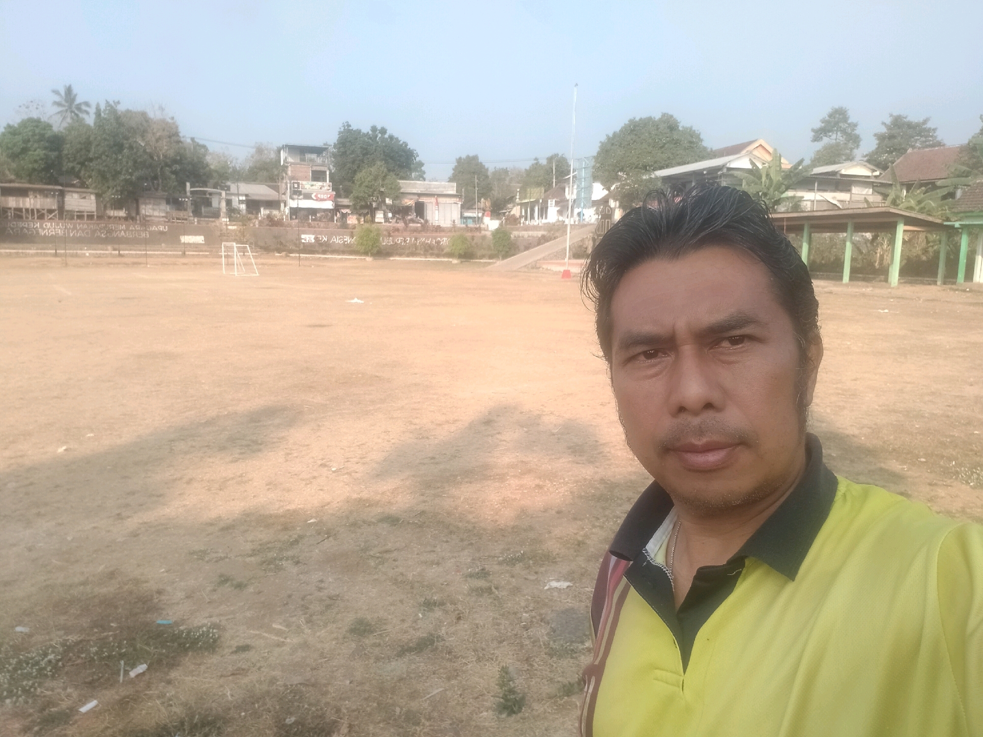 A man is taking a selfie in an outdoor area. The man is in the foreground, facing the camera. He has dark hair, brown eyes, and is wearing a yellow polo shirt with a dark collar and stripes on the sleeves. Behind him is a large, open field that appears to be a sports field, possibly for soccer, with a small white goal visible in the distance. The field is mostly dry and bare, with some patches of grass and scattered debris. In the background, there are several buildings and trees. The buildings appear to be a mix of residential and commercial structures, with some of them looking older. The sky is hazy and light, suggesting it might be early morning or late afternoon. The overall environment suggests a rural or semi-urban setting. The location is identified as the city of Sawahan, Indonesia.