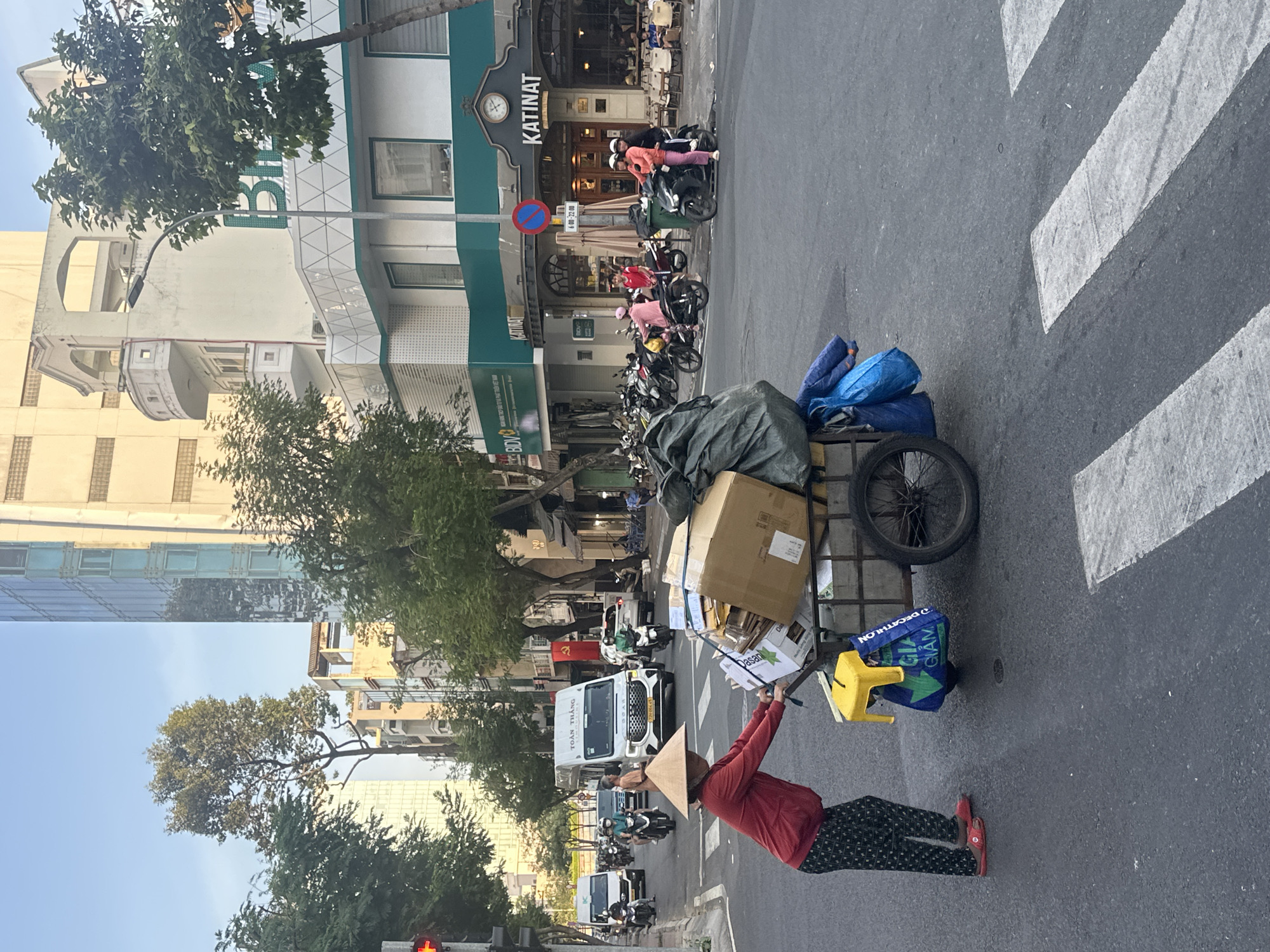 A person wearing a conical straw hat (nón lá), a red long-sleeved shirt, patterned dark pants, and red sandals pushes a metal-framed two-wheeled utility cart laden with cardboard boxes, a large dark green sack, and blue bags, one bearing the "Decathlon Gia Giảm" logo. A yellow plastic stool hangs from the cart. The individual is positioned on an asphalt street with white zebra crossing markings.
The scene takes place on a city street in Thủ Đức, Vietnam, under a clear blue sky. In the background, traffic comprising numerous motorbikes, a white "Toàn Thắng Limousine" van, and other vehicles moves along the multi-lane road. A red traffic light is visible on the left. The right side of the street is lined with multi-story buildings. The ground level features a "Katinat" coffee shop with outdoor seating and a "BIDV" (Bank for Investment and Development of Vietnam) sign. A street light pole and a red-circular "No Stopping/Parking" sign are present. Further down the street on the left, a Vietnamese flag (red with a yellow star) is visible. Trees are interspersed along the street.