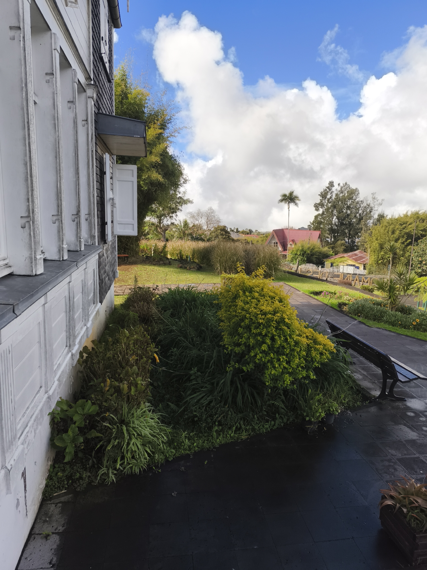 The image depicts a view of a residential area on a partly cloudy day. In the foreground, a white building with louvered shutters and a dark gray roof is visible on the left side of the frame. A paved walkway extends from the foreground towards the middle ground, where a black park bench is situated. Lush green vegetation, including a vibrant yellow-green bush, lines the walkway and extends into the middle ground. Beyond the vegetation, a grassy lawn leads to a street or pathway with a white picket fence. In the background, two houses with red and yellow roofs are visible, surrounded by trees and foliage, including a prominent palm tree. The sky is a clear blue with fluffy white clouds. The overall scene suggests a well-maintained residential environment with a focus on nature and outdoor spaces. The location is identified as La Plaine-des-Palmistes, France.