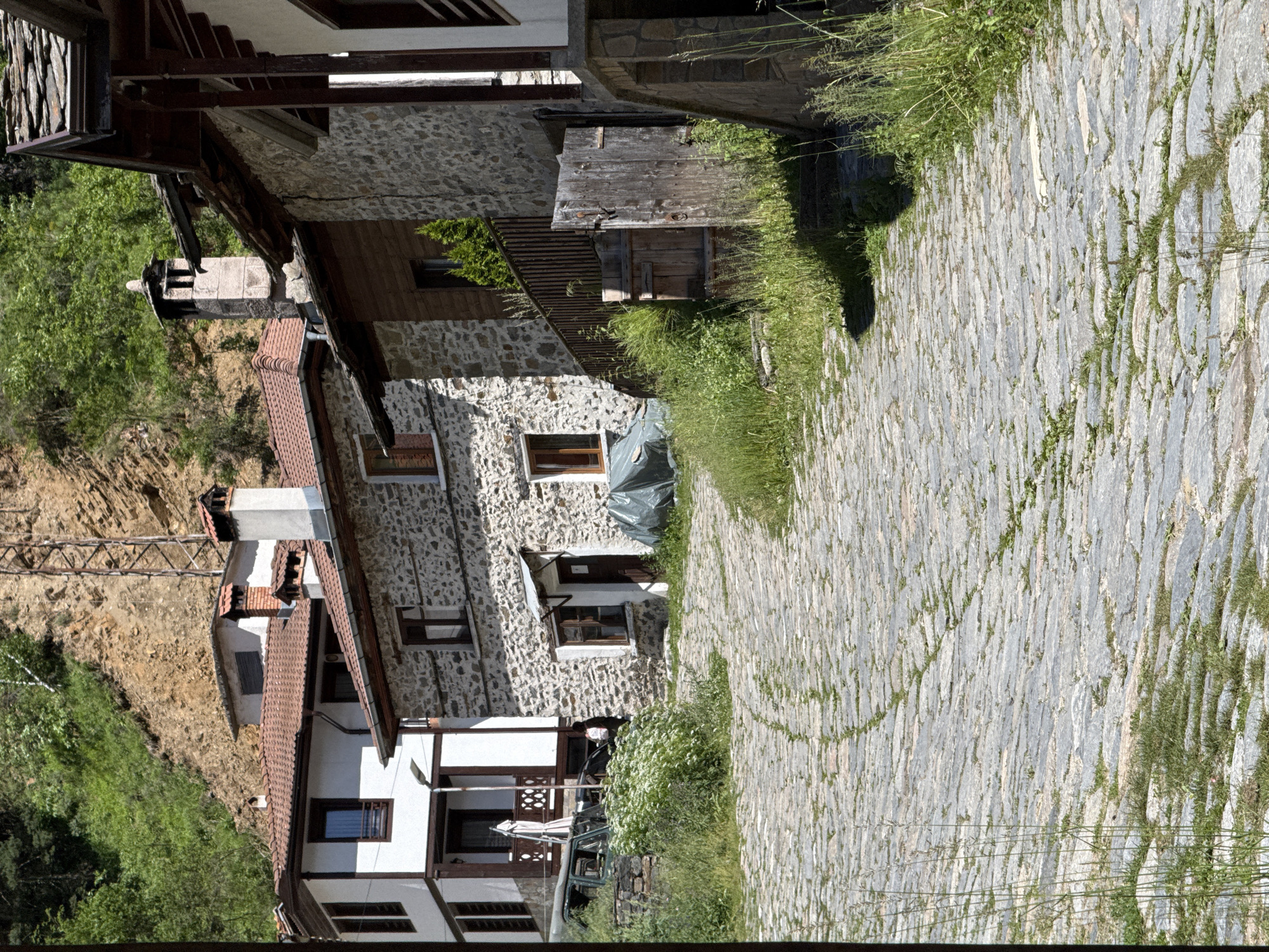 The image captures a cobblestone street winding uphill through a village. On the right, a stone and wooden house with a dark wooden door and a pitched roof is visible, with tall grass growing around its base. To the left of the street, a two-story white house with dark trim and a red tiled roof stands, with a white balcony on its upper floor and a car parked in front. Further up the street, another stone house with a red tiled roof and a prominent brick chimney is seen. The background consists of a steep, rocky hillside covered in green trees and vegetation, with a metal pylon visible on the slope. The scene appears to be in Shiroka Laka, Bulgaria, known for its traditional Rhodope architecture.