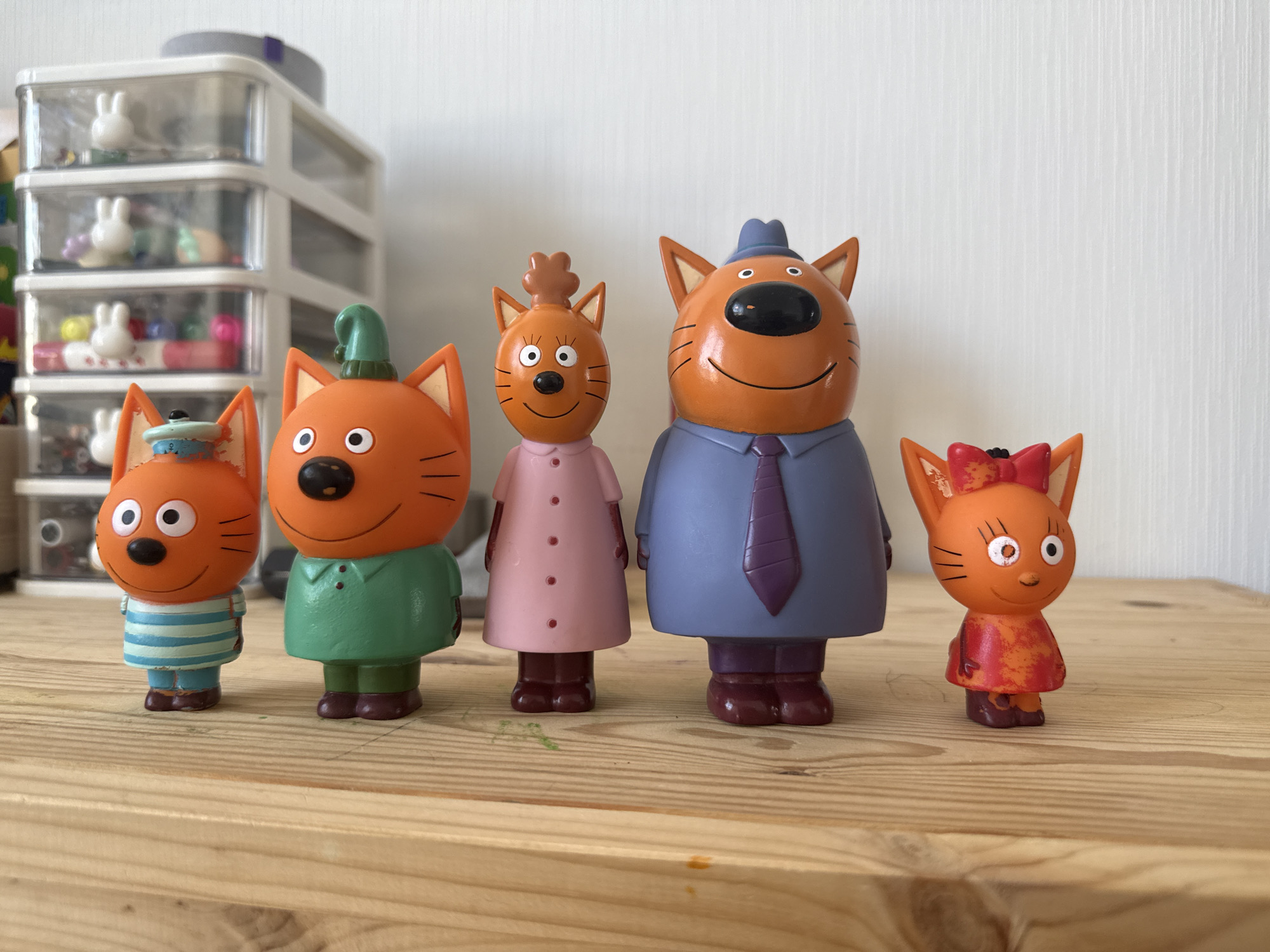 This is a photograph of five orange cat figurines posed on a wooden surface against a white, textured background. The figurines are arranged in a line from left to right. The first figurine on the left is wearing a blue and white striped shirt and a small blue hat with a light blue rim and a white brim. It has large white eyes, a black nose, and thin black lines representing whiskers. The second figurine is wearing a green hooded outfit and has a similar facial expression to the first. The third figurine is wearing a pink dress with four red buttons down the front and has a tuft of hair on its head. The fourth figurine, the largest, is wearing a blue suit jacket, a purple tie, and a blue fedora hat. The fifth figurine, on the far right, is wearing a red and orange patterned dress and a red bow on its head. Behind the figurines, to the left, is a clear plastic organizer with multiple drawers, containing various small items. The wooden surface appears to be a table or shelf made of light-colored planks. The scene is well-lit, with the light source appearing to be from the front and slightly to the left. The photograph is taken from a slightly low angle, looking up at the figurines. There is no information in the image to suggest a specific location in Lyubertsy, Russia.