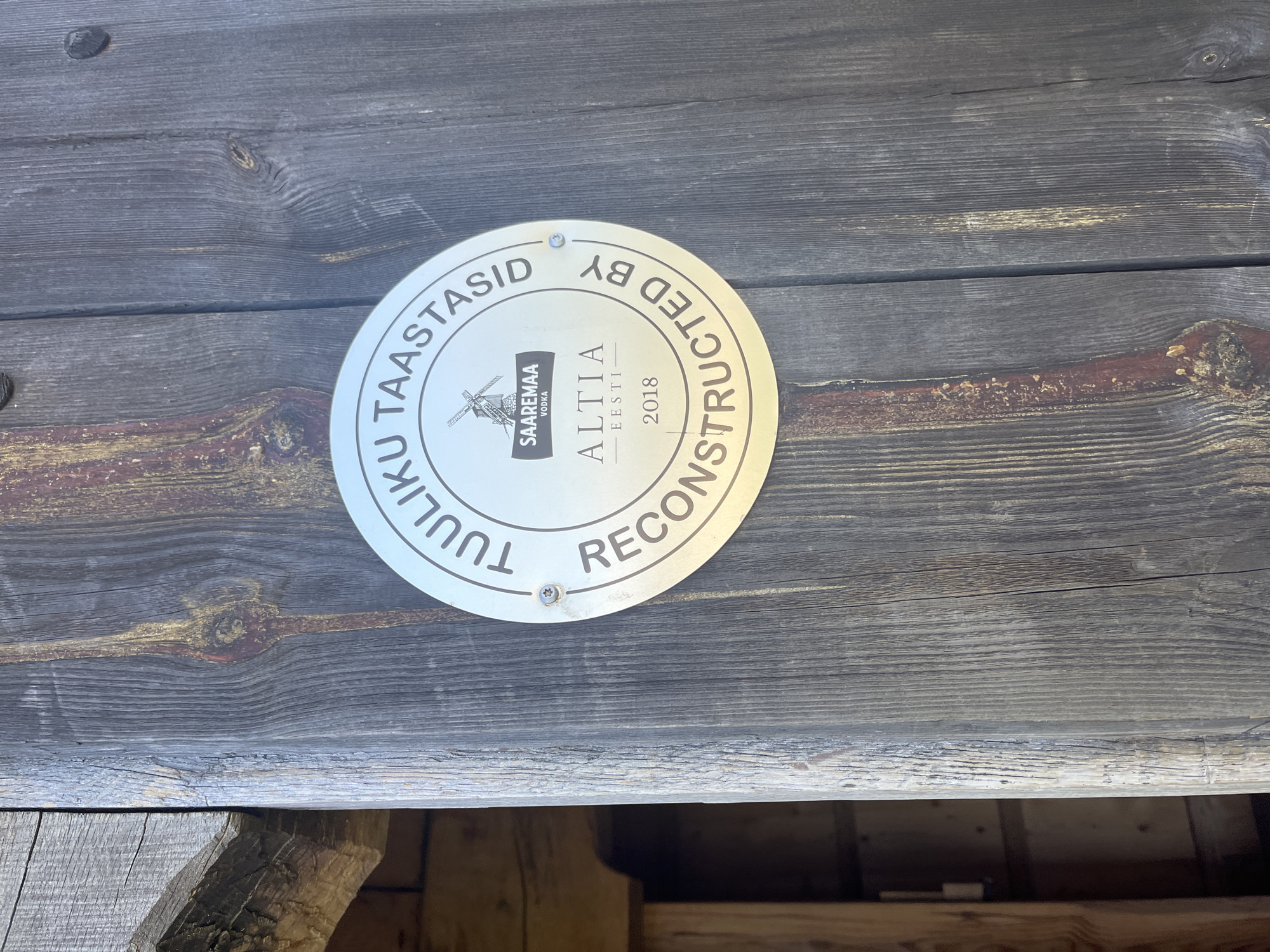 A circular metal plaque is affixed to a weathered wooden door. The plaque features text in an arc along the top and bottom, with a central design. The top arc reads "TUULIKU TAARAASID", and the bottom arc reads "RECONSTRUCTED BY". In the center of the plaque is a windmill icon above the text "SAAREMAA VODKA", followed by "ALTIA EESTI" and the year "2018". The plaque is attached to the door with visible screws. The door itself is made of vertical wooden planks, showing signs of age and texture. To the left of the door, part of a wooden structure is visible, suggesting an entranceway or building. The overall impression is one of historical preservation or restoration.