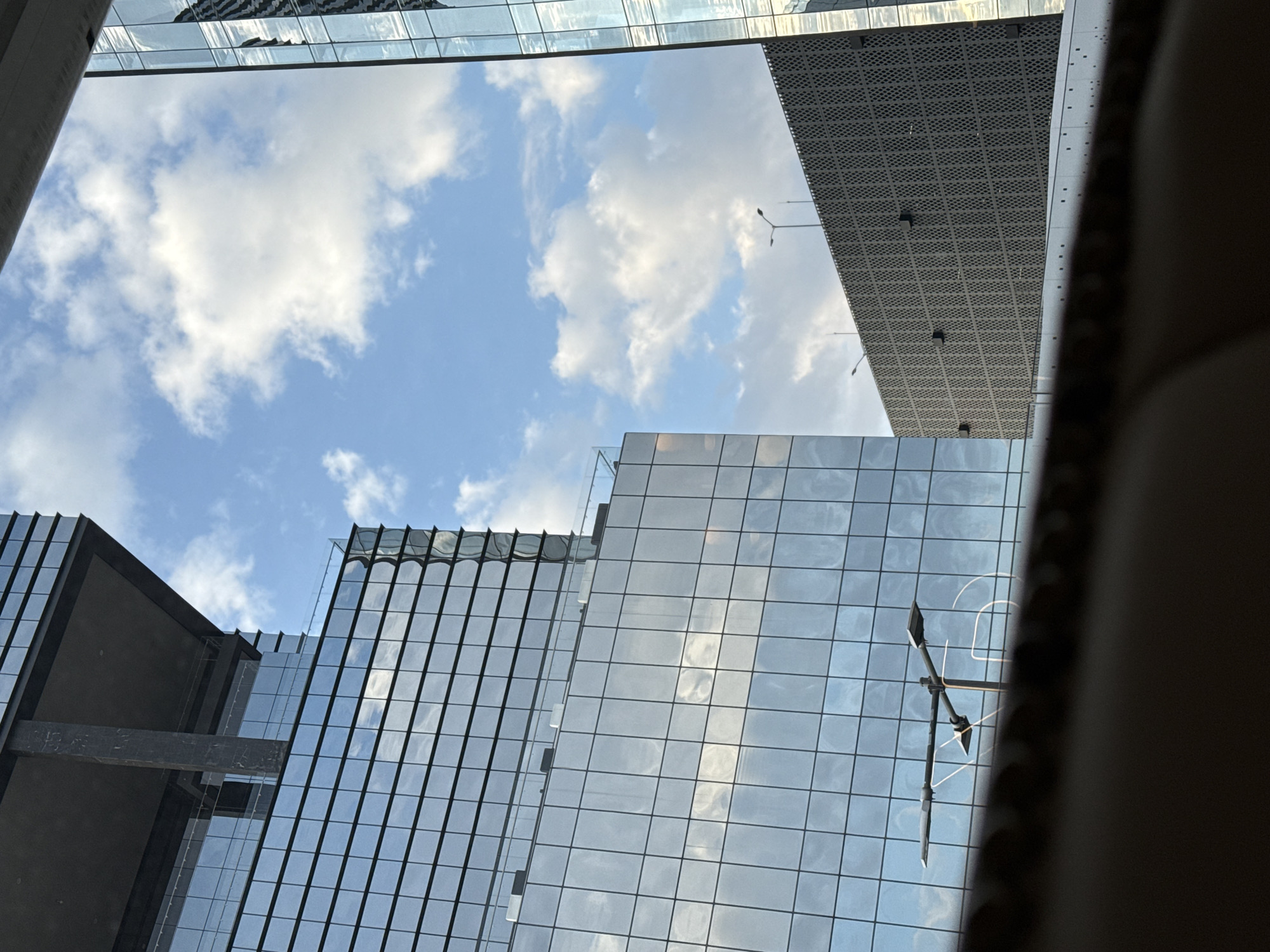 The image presents an upward-looking view of modern urban architecture against a bright blue sky with scattered white, cumulus clouds. The scene is characterized by several tall buildings, predominantly featuring sleek glass facades that reflect the sky, clouds, and surrounding structures, creating intricate patterns of light and shadow.
One prominent building section towards the top right exhibits a dark, perforated paneling, possibly made of metal, offering a textural contrast to the smooth, reflective glass. Streetlights are visible against the sky and as reflections on the glass. On the left side, a darker building with a substantial horizontal beam connecting its sections is seen. The overall aesthetic is one of contemporary design with strong geometric lines.
The clear sky and bright illumination suggest it is daytime, likely late morning or early afternoon, with sunny weather. The perspective, looking up from below, emphasizes the height and imposing nature of the structures. A dark, out-of-focus element partially frames the image on the right and top left, hinting at the photograph being taken from a specific vantage point, possibly inside a vehicle or from a low architectural feature. There are no people visible, and no specific activities are taking place. No text is discernible within the image. The setting aligns with a modern urban center, such as Puebla, Mexico.