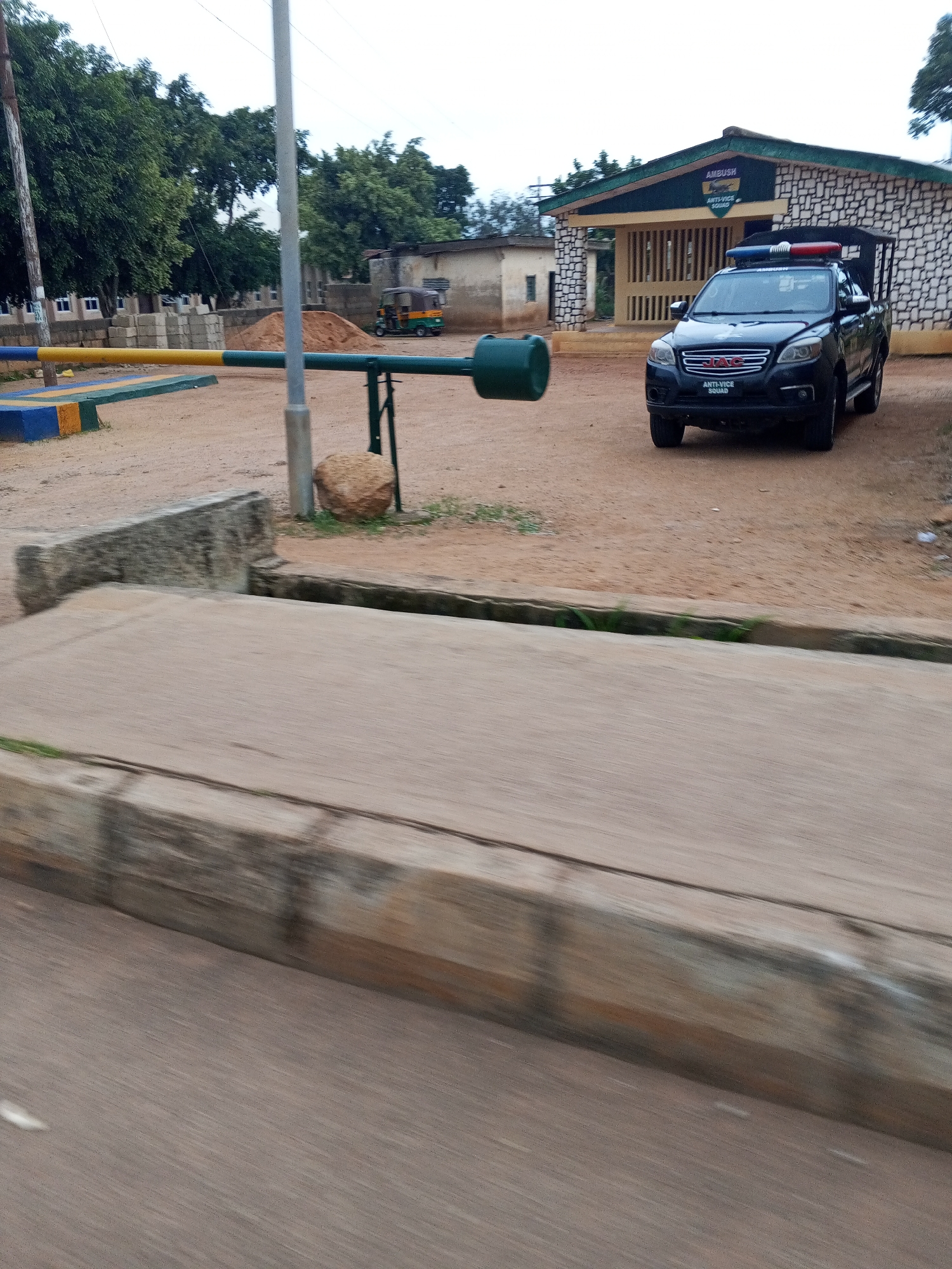 In Bukuru, Nigeria, a black SUV with the words "ANTI-VICE SQUAD" and "AMBUSH" emblazoned on it is parked in front of a building with a stone facade and a green roof. The vehicle is equipped with a light bar on its roof. A yellow and blue striped barrier is positioned across a dirt road, with a green pole extending from it. In the background, trees and a small, colorful tuk-tuk are visible. The overall scene suggests a security checkpoint or outpost.