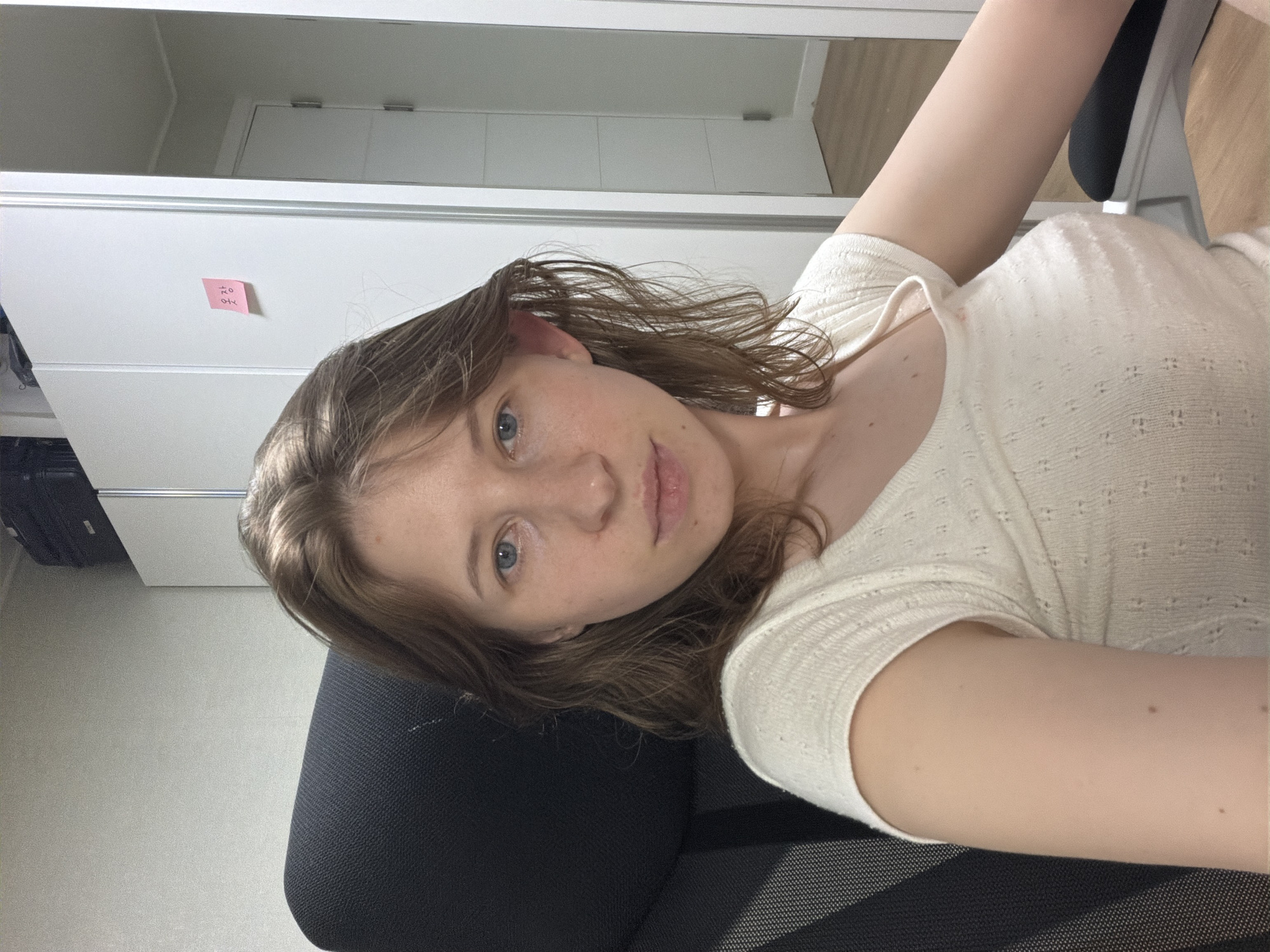 A young woman with light brown hair and blue eyes is sitting in a black office chair, looking directly at the camera. She is wearing a light-colored, short-sleeved knit top. Behind her, to the left, is the back of the office chair. To the right, a white closet door is visible, with a small pink sticky note attached to it that has Korean writing on it. The background appears to be an indoor setting, possibly an office or a room in a residence, with a door and wooden flooring reflected in a mirror or glass surface on the far right. The lighting is natural and soft. The image was taken at an eye-level perspective.