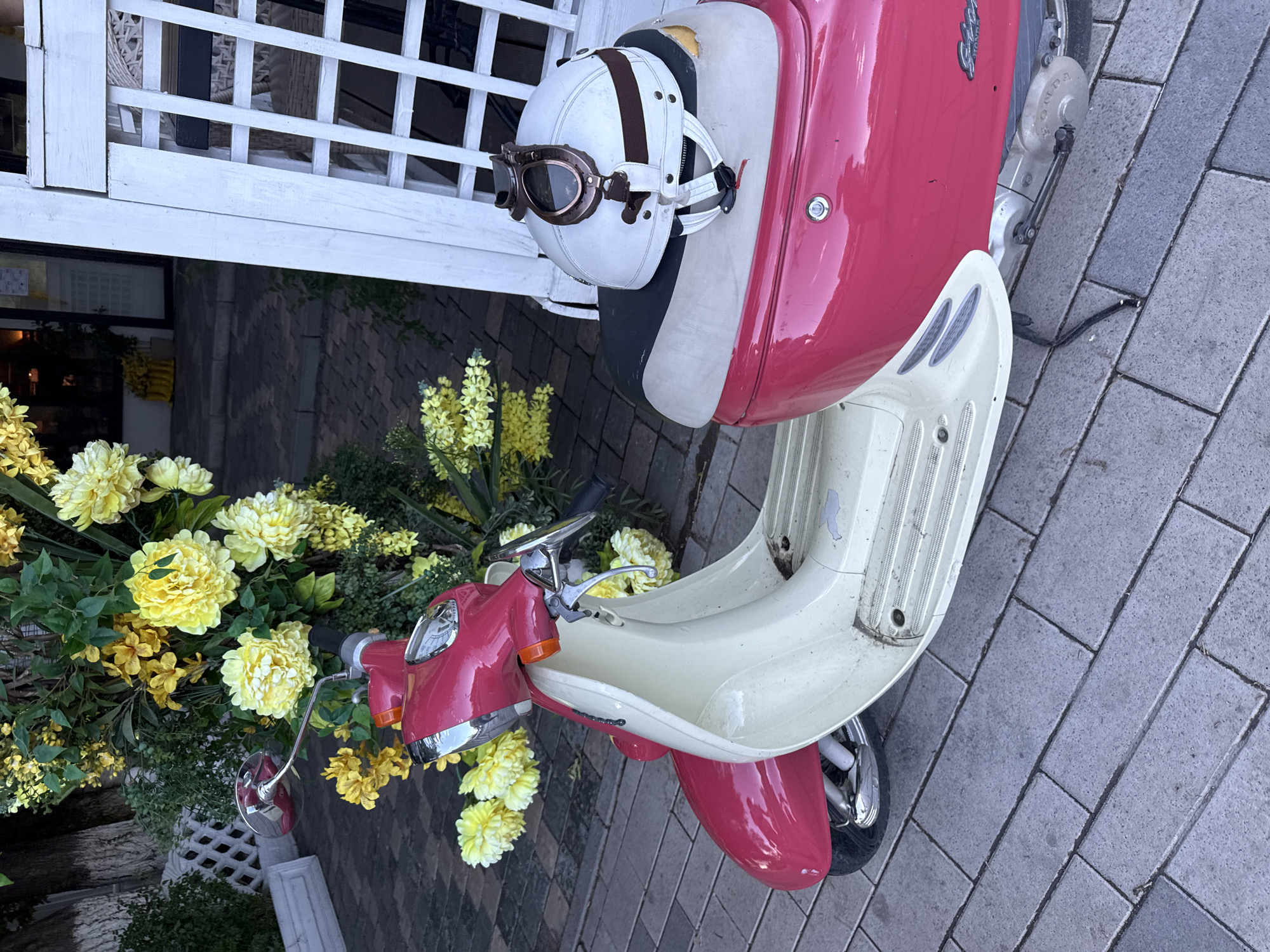 A pink and cream colored scooter is parked on a cobblestone sidewalk in front of a white trellis. A white helmet with goggles is resting on the back of the scooter seat. Behind the scooter, a large floral arrangement with yellow and white flowers is visible. The scene appears to be outdoors, possibly in front of a building with large windows. No people are visible in the image.
