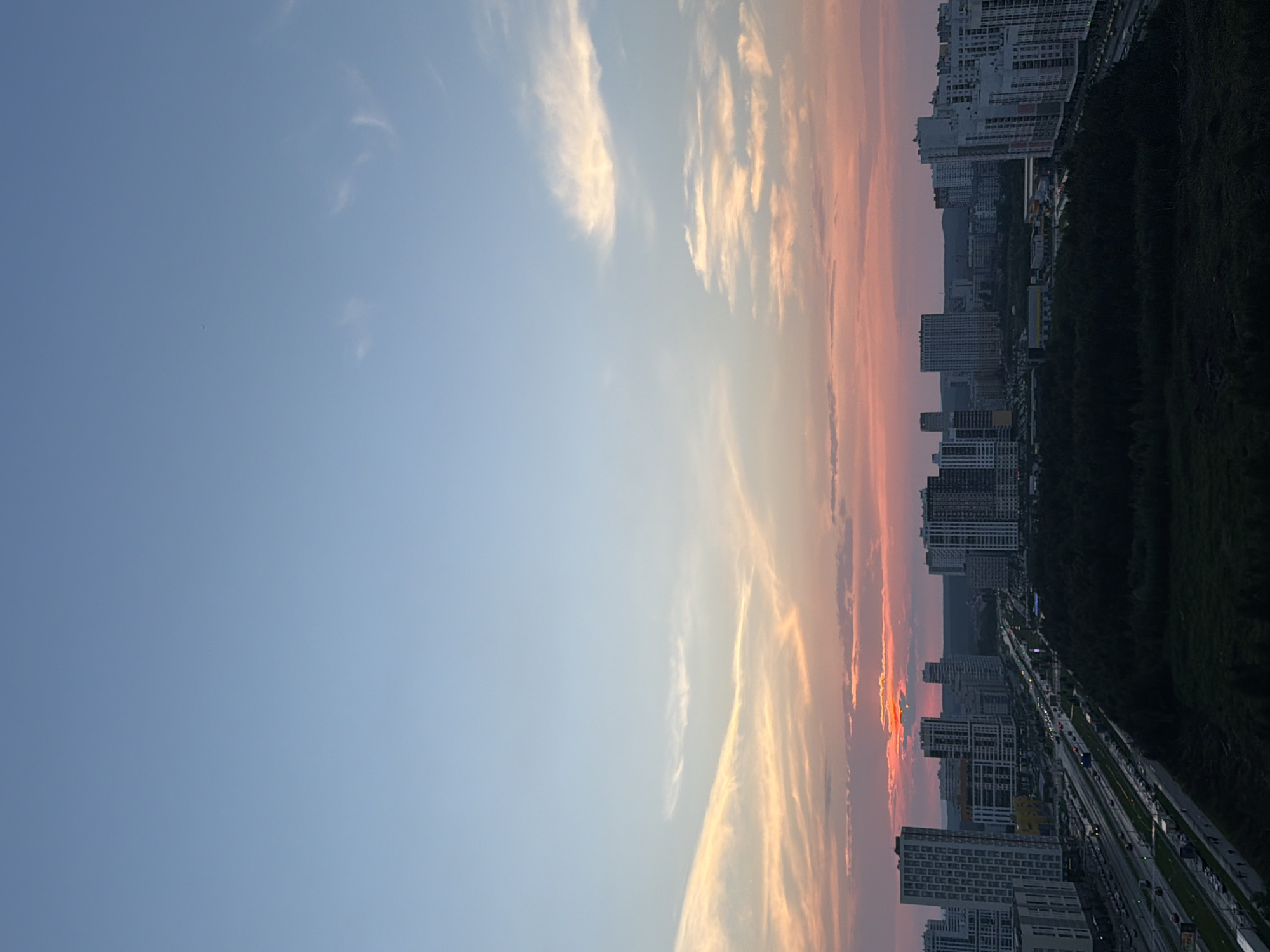 This image depicts a panoramic view of the city of Yekaterinburg, Russia, during sunset. The sky transitions from a clear, light blue at the top to hues of pink, orange, and yellow closer to the horizon, with wispy clouds catching the fading sunlight. Below the sky, a sprawling cityscape unfolds, characterized by numerous modern high-rise buildings, some with white facades and distinct architectural designs. A multi-lane highway runs through the city, with visible traffic, suggesting an active urban environment. Dark green trees form a dense band of forest in the foreground and mid-ground, contrasting with the built environment. The overall scene captures the essence of a city at dusk, with the warm glow of sunset illuminating the buildings and the sky.