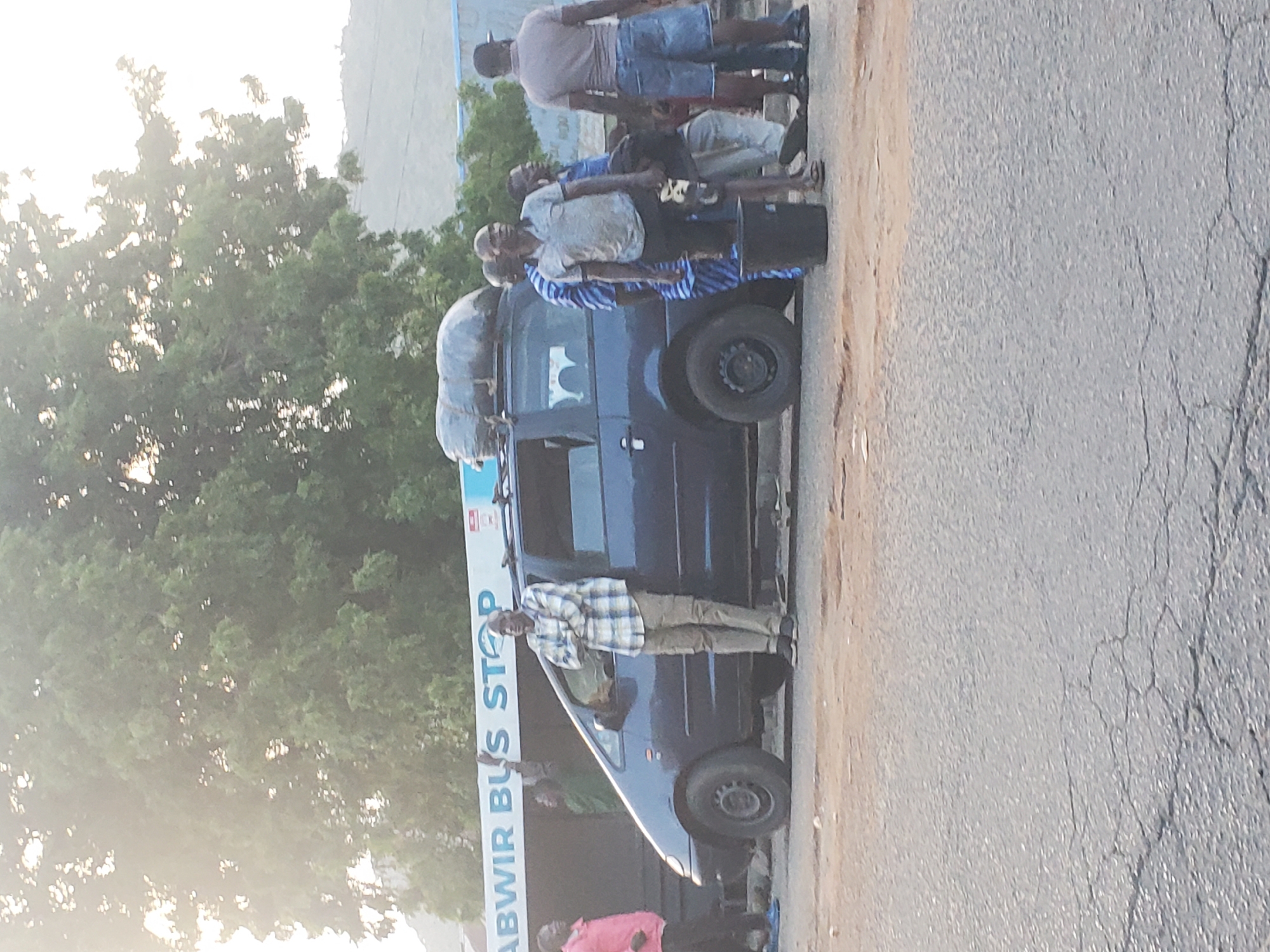 The image shows a group of people standing next to a dark blue van loaded with luggage on its roof. The van appears to be at a bus stop, indicated by a sign that reads "ABWIR BUS STOP". Several individuals are standing around the van, some looking towards the camera, while others seem to be waiting. The people are dressed in casual clothing. One man in the foreground, wearing a plaid shirt, is standing with his arms crossed, looking directly at the camera. The scene is set outdoors, with trees visible in the background and a road in the foreground. The lighting suggests it is daytime, possibly late afternoon, with the sun creating a bright sky. The ground is a mix of dirt and cracked asphalt. The overall impression is one of travel or waiting for transport in a rural or semi-urban setting in Ampan, Nigeria.