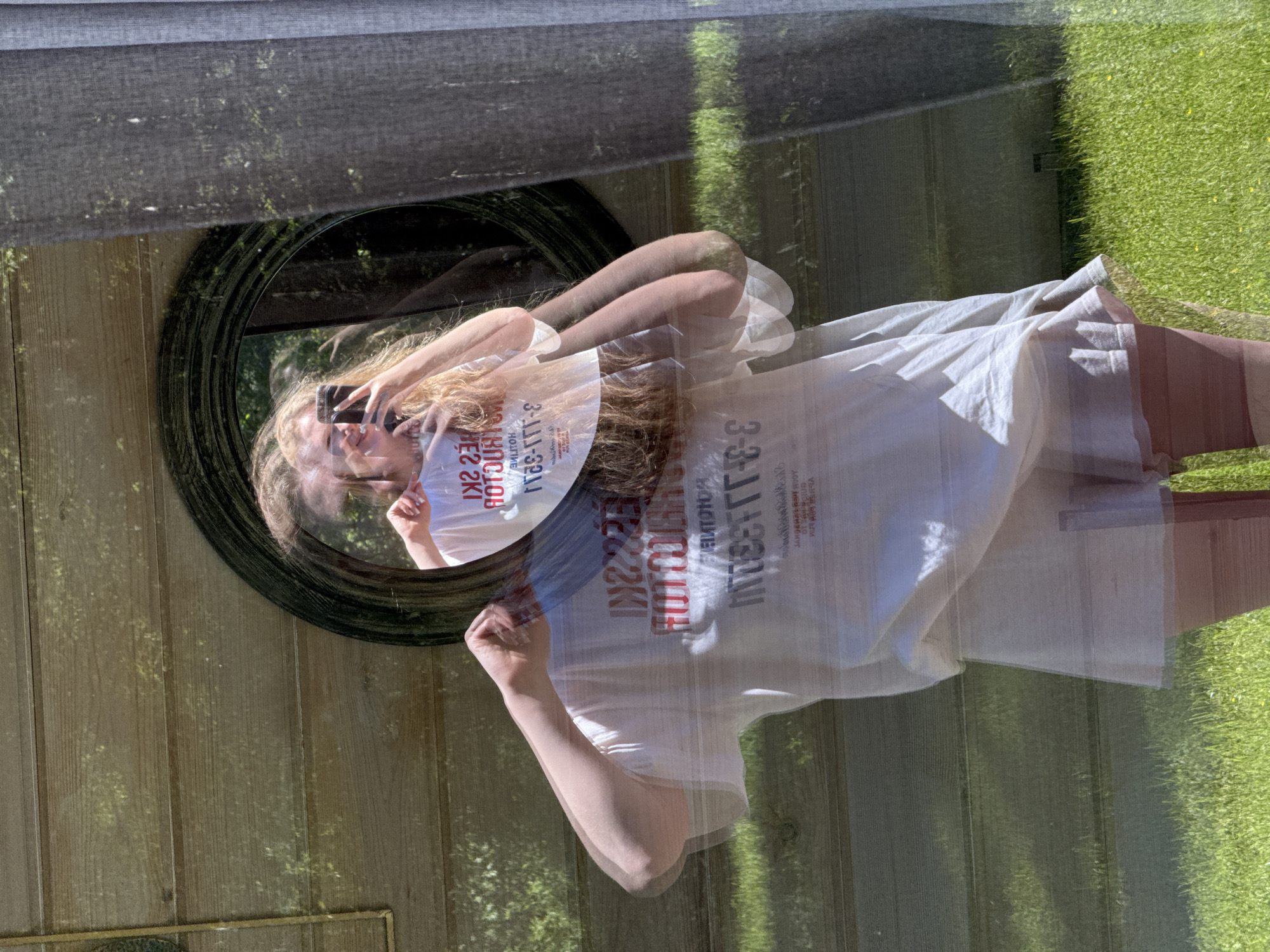 A composite image depicts a mirror selfie captured through a reflective surface, presenting a female subject overlaid on an outdoor scene in Копнино, Russia. The subject, a female with long, light-colored hair, is positioned centrally, facing slightly left, taking a photograph of her reflection in a circular mirror with a dark, ornate frame. She holds a dark-colored smartphone to capture the image. She wears an oversized white t-shirt with red and black text, reversed in the mirror to read "SKI HOTLINE 3-111-S2H" and appearing as a translucent overlay on her form. Light-colored shorts are visible beneath the t-shirt, along with her legs, which appear to be covered by light-colored hosiery or leggings. The subject in the mirror reflection exhibits a smile. The background, visible through the subject's form and around the mirror, shows a brightly lit outdoor environment with a horizontally slatted wooden fence or wall on the left, vibrant green grass in the lower half, and a dark grey, pleated curtain on the right. Dappled sunlight suggests an overhead tree canopy, illuminating the wooden wall and grass.
