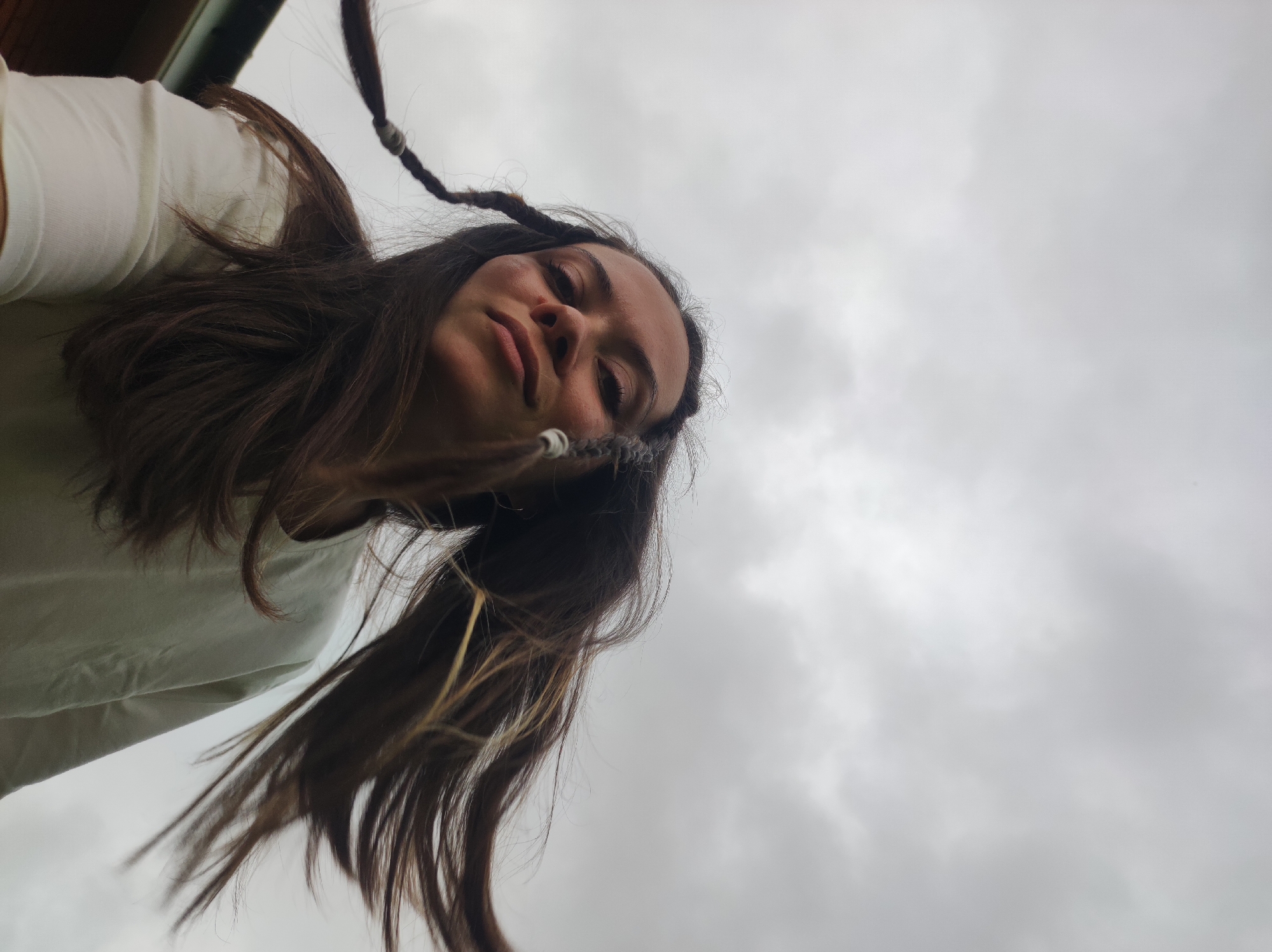 The image portrays a young adult female from a low-angle perspective, looking downwards towards the camera, against an overcast sky in Foki, Russia. She has long, dark brown hair, with several braids or dreadlocks visibly adorned with light-colored, possibly white or grey, wraps or bands. Her eyes are closed or partially closed, and her facial expression appears serene or subtly smiling. She is wearing a light-colored, long-sleeved top. The background consists predominantly of a diffused, grey cloudy sky. A dark, narrow, horizontal structure or element is partially visible in the extreme upper-left corner of the frame, suggesting an immediate overhead environment. The lighting is soft and even, consistent with overcast conditions.