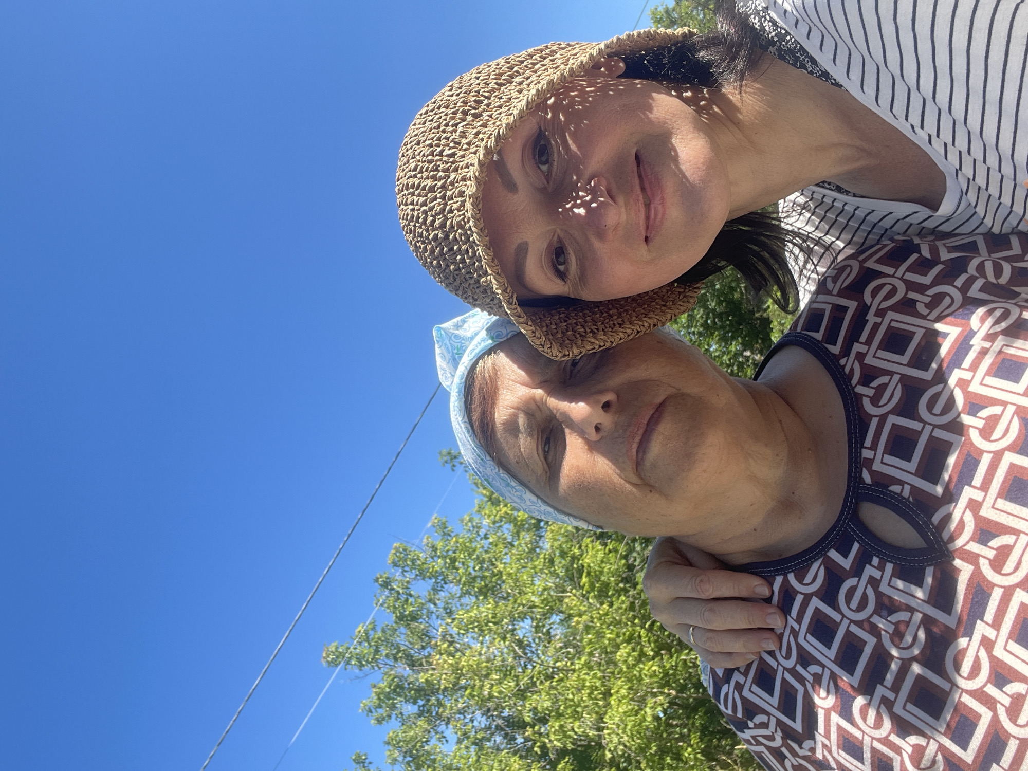 This is a selfie taken outdoors on a sunny day with a clear blue sky. Two women are pictured from the chest up, with a leafy green tree and power lines visible in the background. The woman on the left, who appears older, is wearing a patterned top and a light blue headscarf. Her left arm is around the shoulders of the woman on the right. The woman on the right, who appears younger, is wearing a straw hat and a striped shirt. The image appears to have been taken in or near the city of Дресвяново, Russia, based on the context provided.