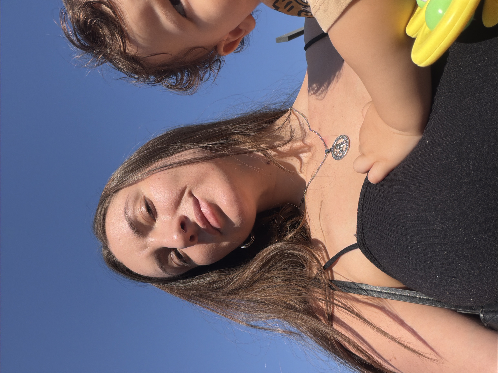 A young woman with long brown hair is holding a baby against her chest in front of a bright blue sky, suggesting an outdoor setting. The woman is wearing a black camisole and a silver pendant necklace. The baby's face is partially visible on the right side of the image, with curly brown hair. The baby's hand rests on the woman's chest, and a yellow toy is visible in the lower right corner. The lighting indicates a sunny day. The scene appears to be located in the city of Dahab, Egypt, given the context of the location.