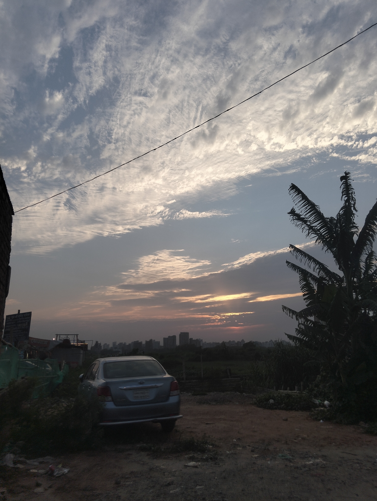 The image depicts a silver sedan parked on a dirt road in Dhaka, Bangladesh, with a view of the skyline at sunset. The sky is filled with textured clouds, and a power line stretches across the upper part of the frame. A banana tree is visible to the right. The car is surrounded by overgrown vegetation and trash. In the background, buildings can be seen silhouetted against the setting sun. The scene appears to be located in a less developed area of the city.