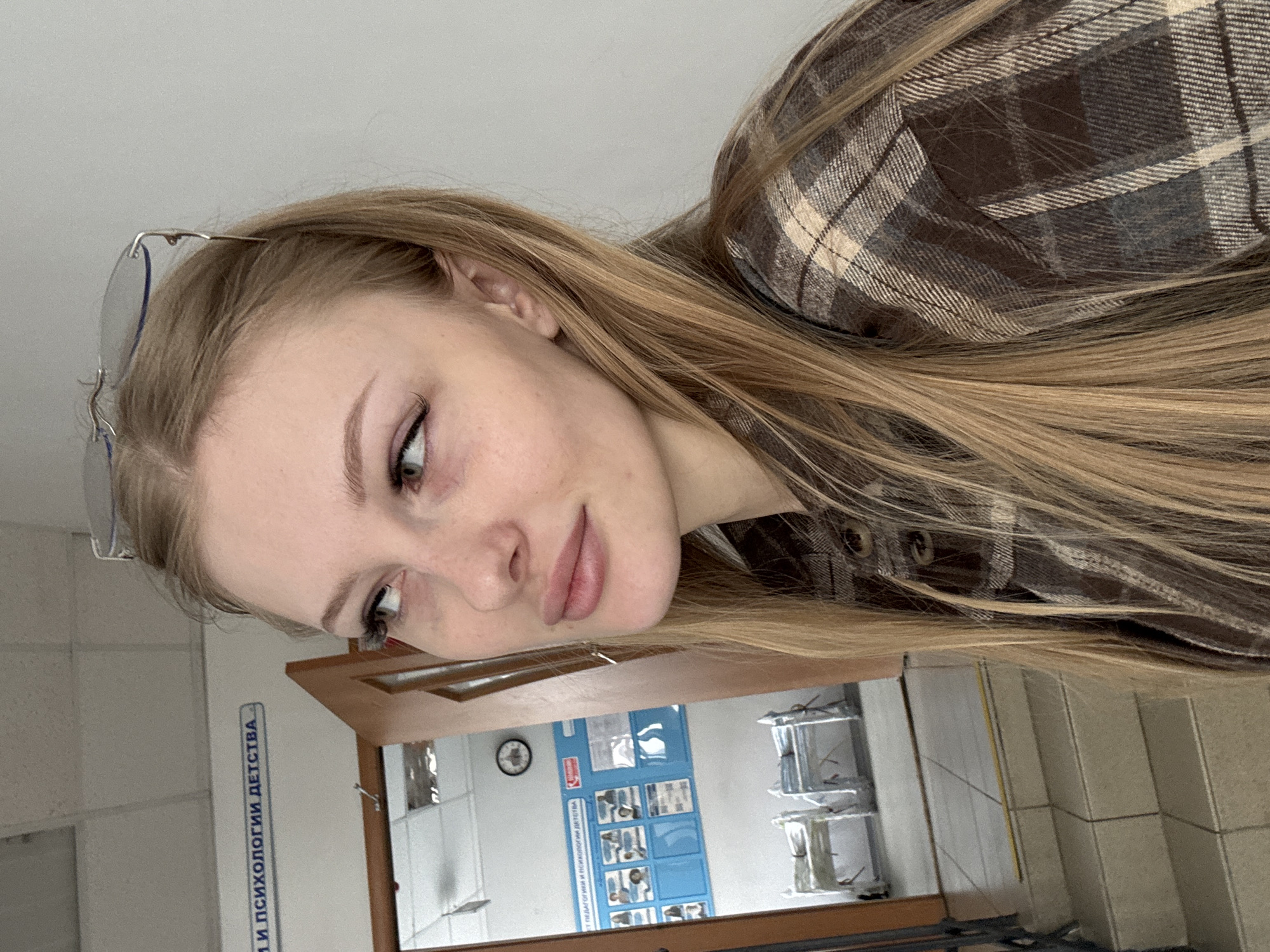 The media file is a portrait of a young woman with blonde hair and light skin, wearing a brown and white plaid shirt. She is looking towards the camera with a slightly tilted head and a gentle smile. Her eyes are light green and she is wearing subtle eye makeup. She also has a smattering of light freckles across her nose and cheeks. She is wearing glasses that are resting on her head.
The background appears to be an indoor setting, possibly a classroom or a laboratory, given the presence of a wooden cabinet with multiple shelves and some posters or charts displayed on the wall. One poster has blue backgrounds and appears to contain images and text in Russian. A clock is visible on the wall. There is also a blue sign with text in Russian. The overall lighting suggests it is daytime.
The sign on the wall reads: "И ПСИХОЛОГИИ ДЕТСТВА" which translates to "AND CHILDHOOD PSYCHOLOGY".
