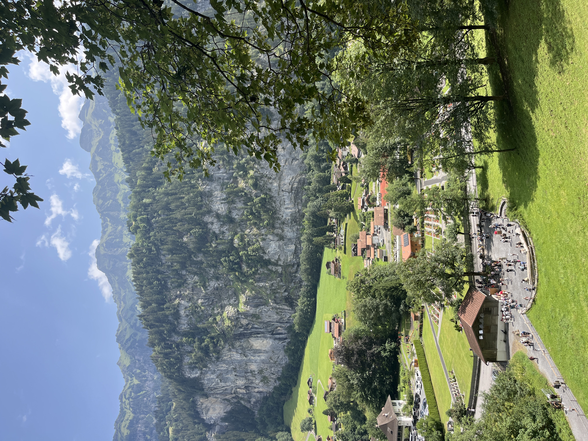 The image captures a scenic view of the Swiss Alps, likely from a high vantage point, overlooking a village nestled in a valley. The foreground is dominated by lush green grass and trees, with dappled sunlight casting shadows. In the middle ground, a cluster of traditional Swiss chalets with red-tiled roofs are spread across verdant fields, bordered by a sheer, imposing rock face covered in dense evergreen trees and a thin waterfall.
In the lower part of the image, a pathway winds through a grassy area, with several people walking and gathered. A small, wooden building with a red roof is situated near a group of people interacting. Further to the left, a parking lot filled with cars is adjacent to a manicured lawn and what appears to be a cemetery with rows of markers.
The background features a dramatic mountain range, with sharp peaks partially obscured by greenery and a clear blue sky with scattered white clouds. The overall impression is one of a serene, natural landscape combined with a well-maintained village, typical of the Bernese Oberland region.