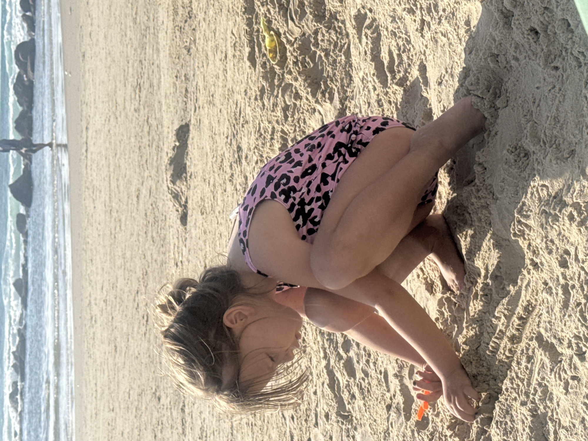 A young girl, wearing a pink and black leopard-print swimsuit, is crouched on the sand at the beach in Bat Yam, Israel. She is facing to the right and digging in the sand with a small orange shovel. The beach is sandy and textured, with visible waves and rock formations in the background, and other people are visible in the distance.