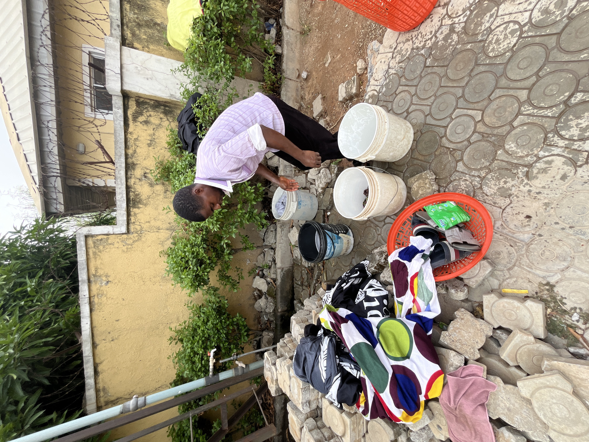 The scene depicts a man outdoors, likely in a residential area of Abuja, Nigeria, engaged in what appears to be hand-washing laundry. He is wearing a light purple, long-sleeved shirt and dark pants, bending over a row of several white plastic buckets filled with water.
To his left are some green plants, growing against a yellow building with a small, barred window visible. A pipe runs along a metal fence in front of the building. Adjacent to the buckets, a pile of colorful laundry sits atop a stack of concrete blocks. An orange basket contains shoes and other items. The ground is paved with patterned concrete tiles, and there is some dirt and gravel present. The overall scene suggests a domestic chore being performed in a typical urban setting.