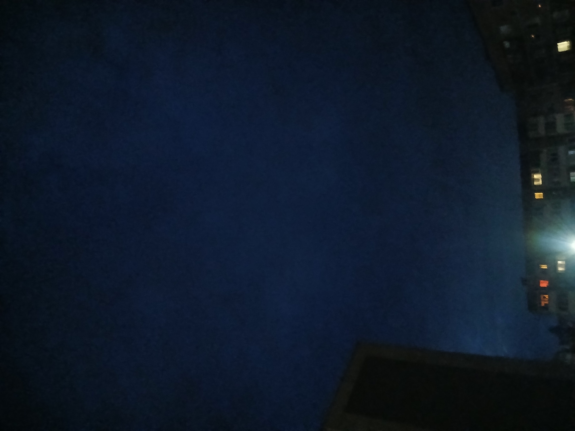 The image is a dimly lit, vertical photograph of a night sky over an urban area, likely Chaikovsky, Russia. The sky is a deep, dark blue, with subtle variations in tone suggesting clouds. In the lower left corner, a portion of a dark, rectangular building is visible, its surface featureless and indistinct due to the low light. To the right and lower, a segment of another apartment building is discernible. Several windows in this building are illuminated with warm, orange-yellow light, indicating occupied residential units. Other windows appear dark. There are bright, white light sources emanating from the lower part of the frame, possibly streetlights or reflections, which create lens flare effects that streak upwards. The overall scene depicts a quiet, nighttime urban landscape.