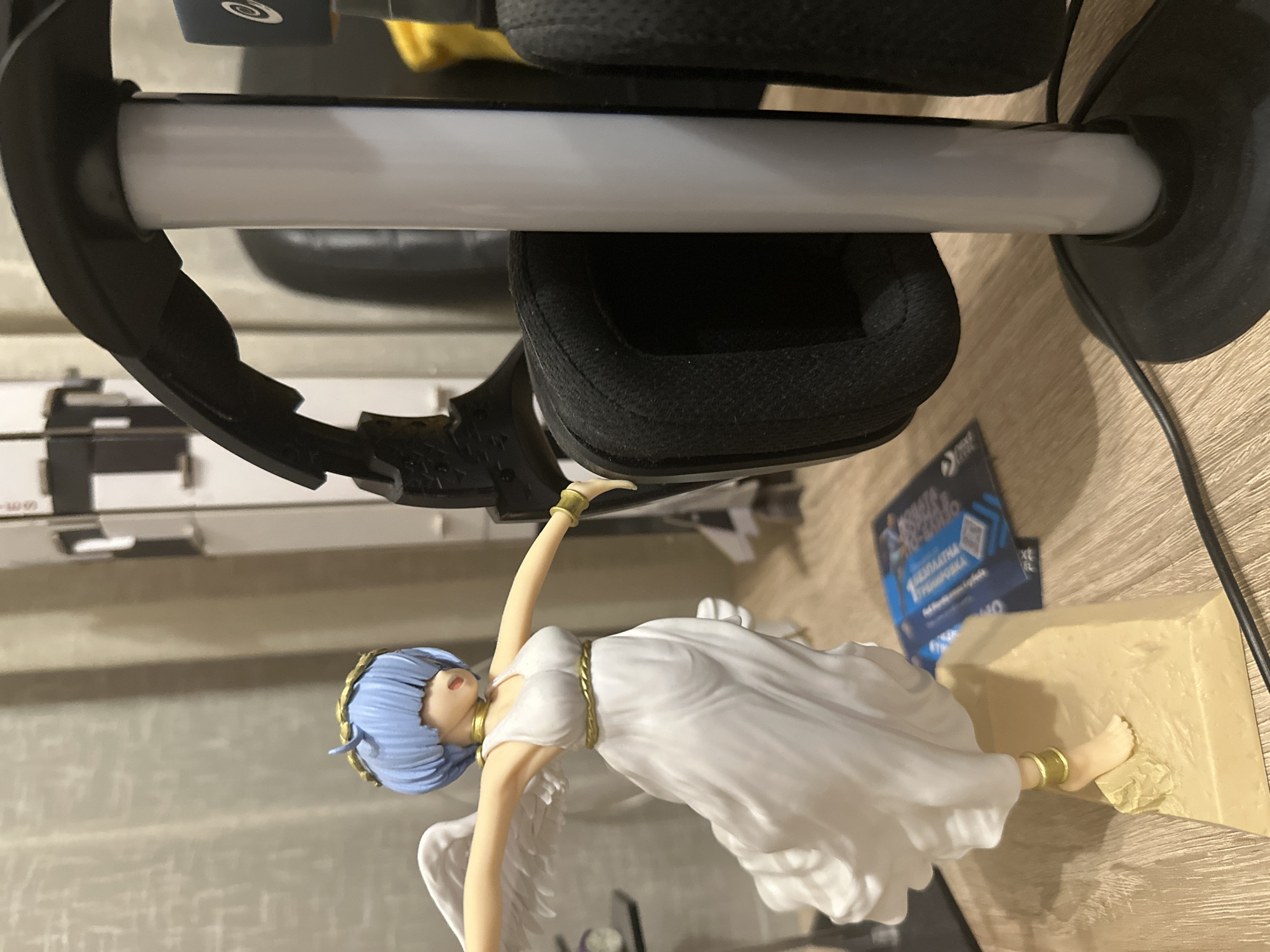 An angel figurine with short blue hair and white wings is standing on a tan platform, with its arms outstretched. It is wearing a flowing white dress with a gold belt and gold armbands. Behind the figurine is a black gaming headset on a white stand. A wooden table and curtains are visible in the background.