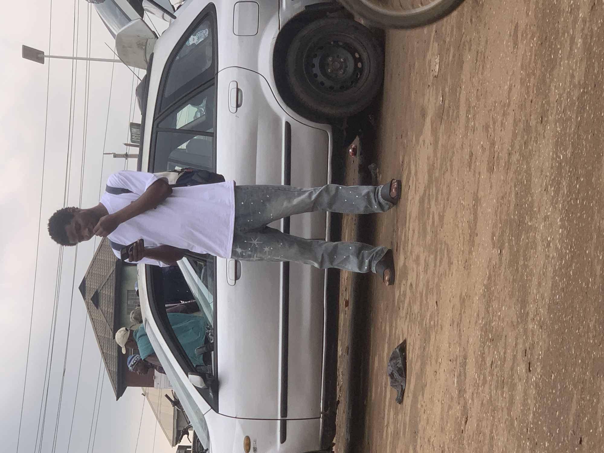 In Shagamu, Nigeria, a young Black male stands on a dirt road next to a silver station wagon, holding a phone to his ear. He is wearing a white t-shirt, light blue jeans with white speckles, and dark sandals. Behind him, other people are visible in and around the car. The car's rear liftgate is open. The ground is dusty and uneven, with a black plastic bag lying on the ground near the man's feet. The sky is overcast.