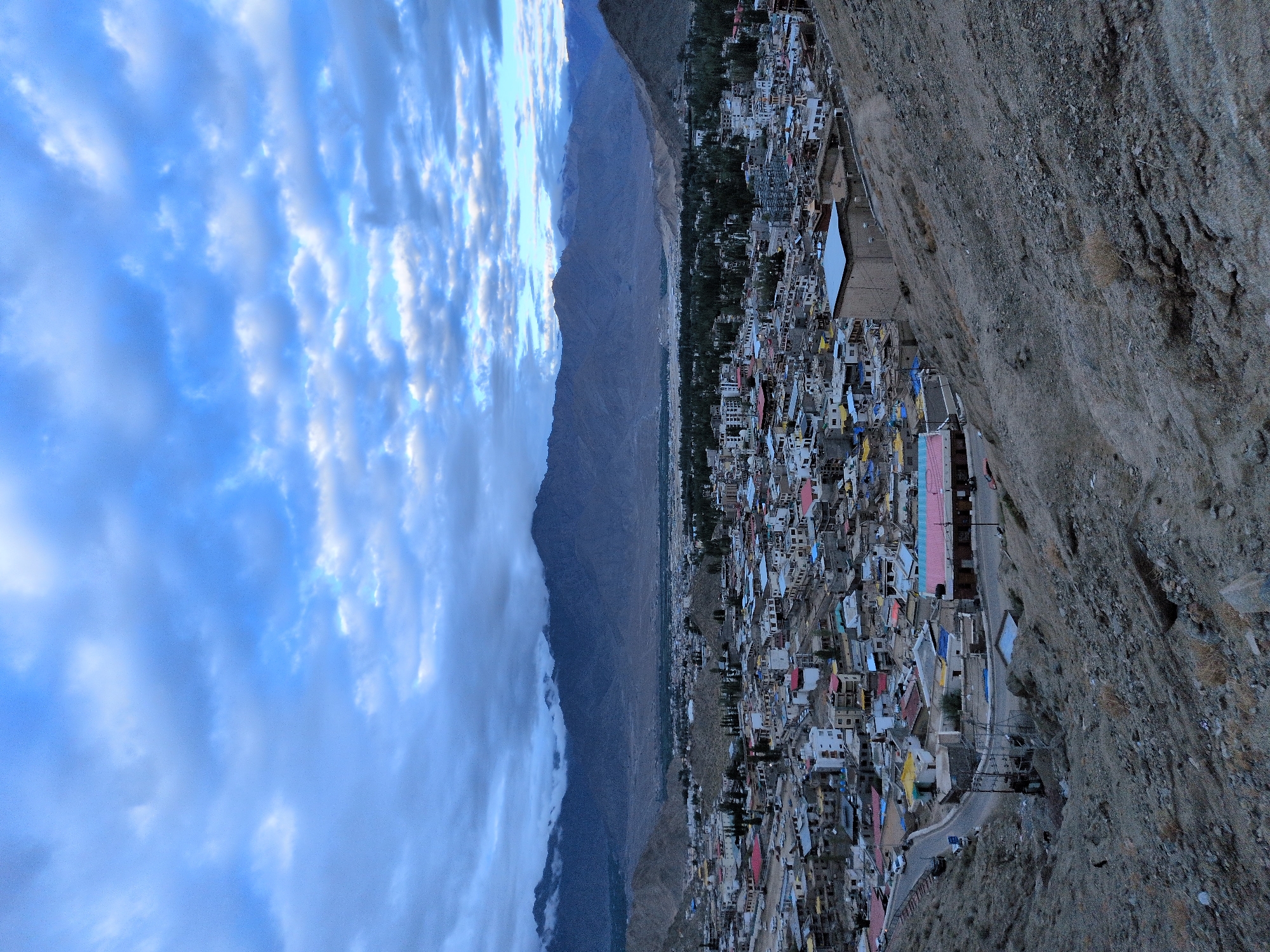 The image depicts an aerial view of the city of Leh, India, at dusk. The scene is dominated by a sprawling urban landscape nestled amidst a mountainous terrain. The city, characterized by a dense collection of buildings with predominantly flat roofs, extends across a valley. The architecture appears to be a mix of traditional and modern, with some structures featuring brightly colored roofs, possibly made of corrugated metal, in shades of red, blue, and yellow. Streets wind through the city, with a few vehicles visible, suggesting active transportation.
The foreground is characterized by a steep, rocky slope covered in sparse, dry vegetation, indicating an arid environment. The middle ground showcases the urban expanse, with individual buildings clearly discernible. In the background, imposing mountain ranges rise, their peaks partially shrouded in clouds, a common feature in the high-altitude region of Ladakh. The sky above is a canvas of blues and grays, with scattered clouds tinged with the fading light of dusk, creating a serene and somewhat dramatic atmosphere. The overall impression is one of a resilient city established within a rugged and majestic natural setting.