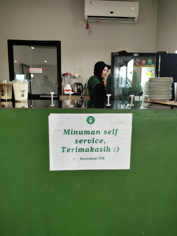 An indoor scene captures the counter area of a coffee shop or cafe, likely in Dramaga, Indonesia. A female barista, wearing a black cap and a green uniform, stands behind a dark counter. To her left, two plastic cups filled with layered beverages are visible. Behind her on the counter are a red blender and a black electric kettle. To her right, a glass-front refrigerator holds various items, and a tall stack of white plates is neatly arranged. An air conditioner is mounted on the light-colored wall above the counter.
In the foreground, a white sign with green text is taped to the dark green counter. The sign reads: "Minuman self service, Terimakasih :)" which translates to "Drinks self service, Thank you :)". Below this, it indicates "Bentokopi IPB", likely the name of the establishment. In the background, behind the counter, a "STAFF ONLY" sign is visible on a glass door, suggesting a restricted area. The reflections of people or objects can be seen in the glass surfaces, indicating a bustling or active environment. The overall setting appears clean and organized, typical of a service area in a commercial establishment.