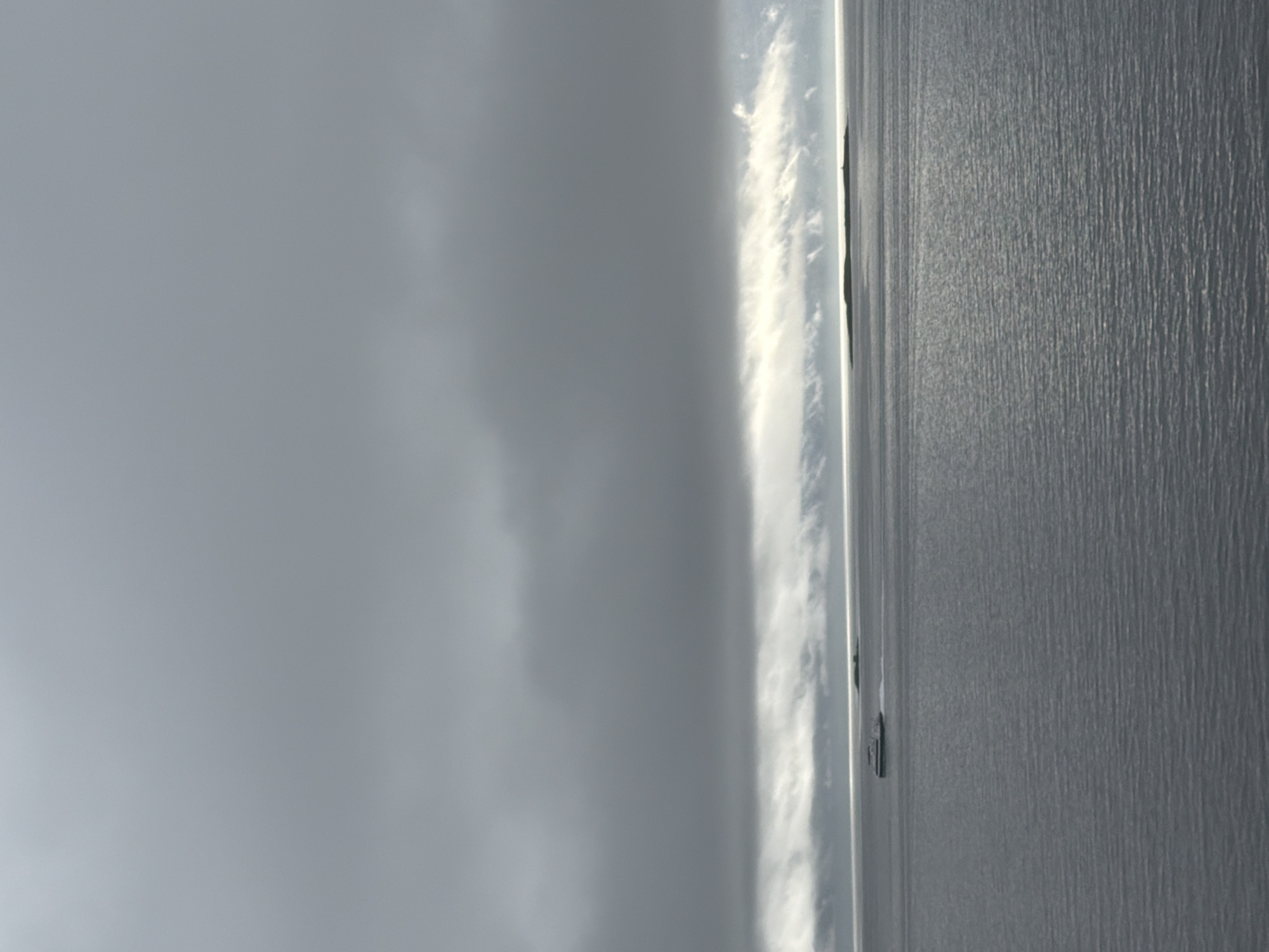 The image shows a vast expanse of calm, dark water under a heavily overcast sky. A band of lighter clouds stretches across the horizon, creating a subtle illumination. In the distance, a long, low-lying island is visible, appearing dark against the brighter sky. A small, white boat with a dark top is moving across the water to the left of the island. The water is textured with gentle ripples. The scene is devoid of any recognizable structures or human activity beyond the single boat. The overall impression is one of a muted and somber seascape. While the image depicts a body of water, it is not possible to determine if it is located in Harstad, Norway, based solely on the visual information provided.