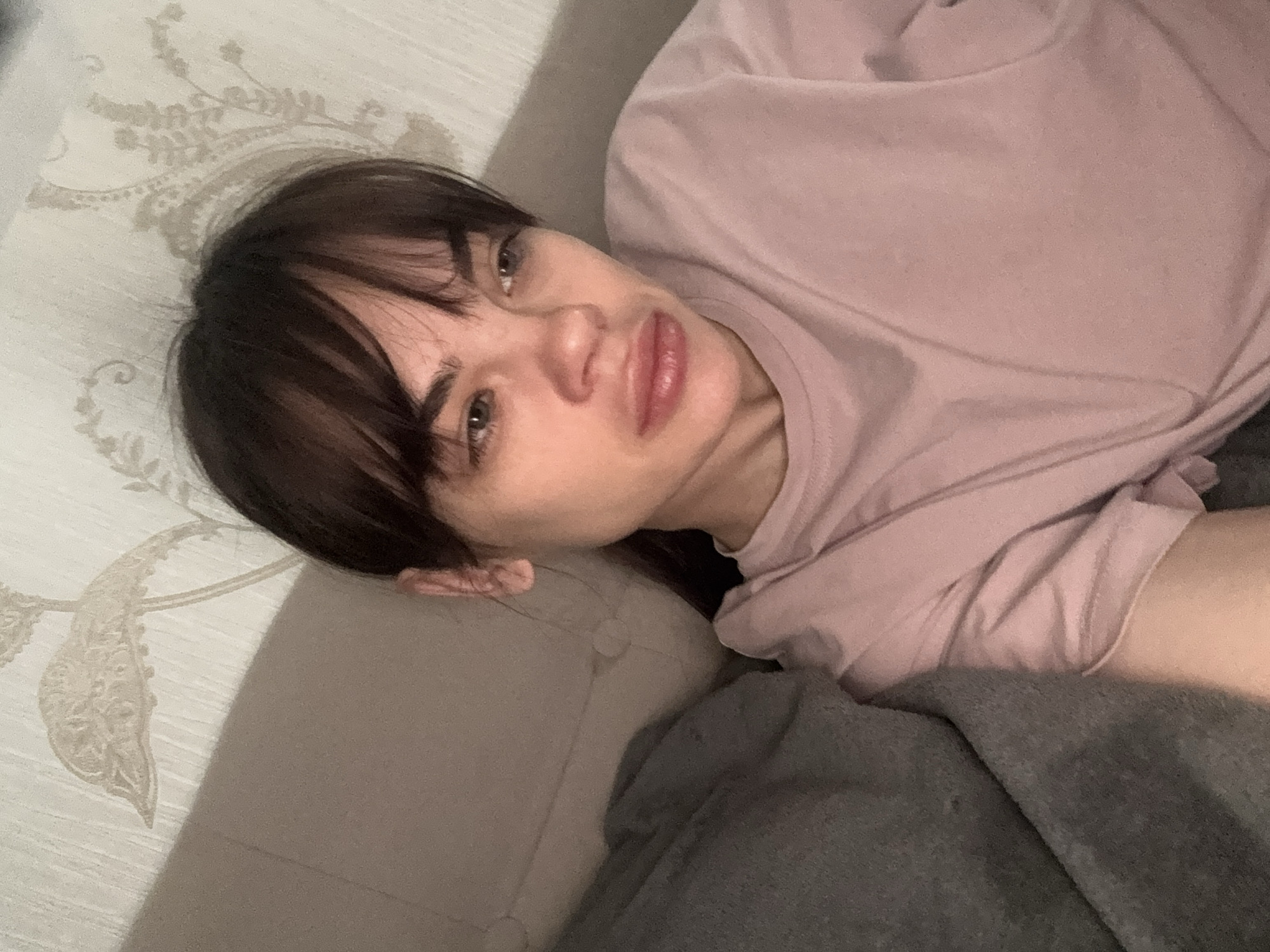 A person with dark brown hair and bangs lies on a sofa or bed. The person is wearing a light pink t-shirt. The background consists of light-colored patterned fabric, possibly a bedsheet or sofa covering, and a grey blanket. The person is looking at the camera. There is no information in the image to indicate the location is Rostov-on-Don, Russia.