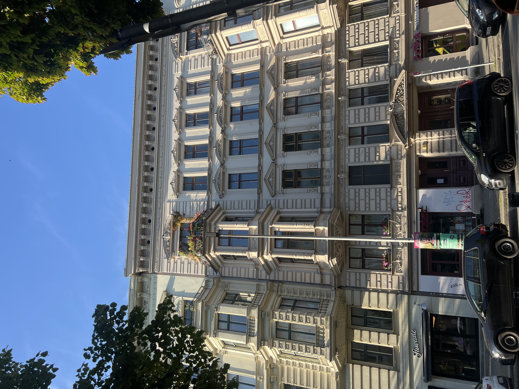 This is a low-angle, eye-level, full shot of a building's facade in Berlin, Germany. The building is constructed in an ornate architectural style, with multiple stories featuring bay windows, decorative cornices, and elaborate window frames. The building is light-colored, likely stone or stucco, and illuminated by sunlight from the right.
On the ground floor, there are storefronts. The leftmost storefront displays a sign that reads "Lawade." To its right, there are entrances with dark wooden doors and a sign with a woman's face and text, appearing to be an advertisement or poster on a pole. Further to the right, another storefront has a sign that reads "ZORA SUPERMARKT".
In front of the building, parked cars are visible along the street. A black hatchback is prominently featured in the foreground, parked on the side of the road. The sky above is a clear, bright blue, with some green tree foliage visible in the upper left and right corners of the frame. The overall impression is of a sunny day in a European city.