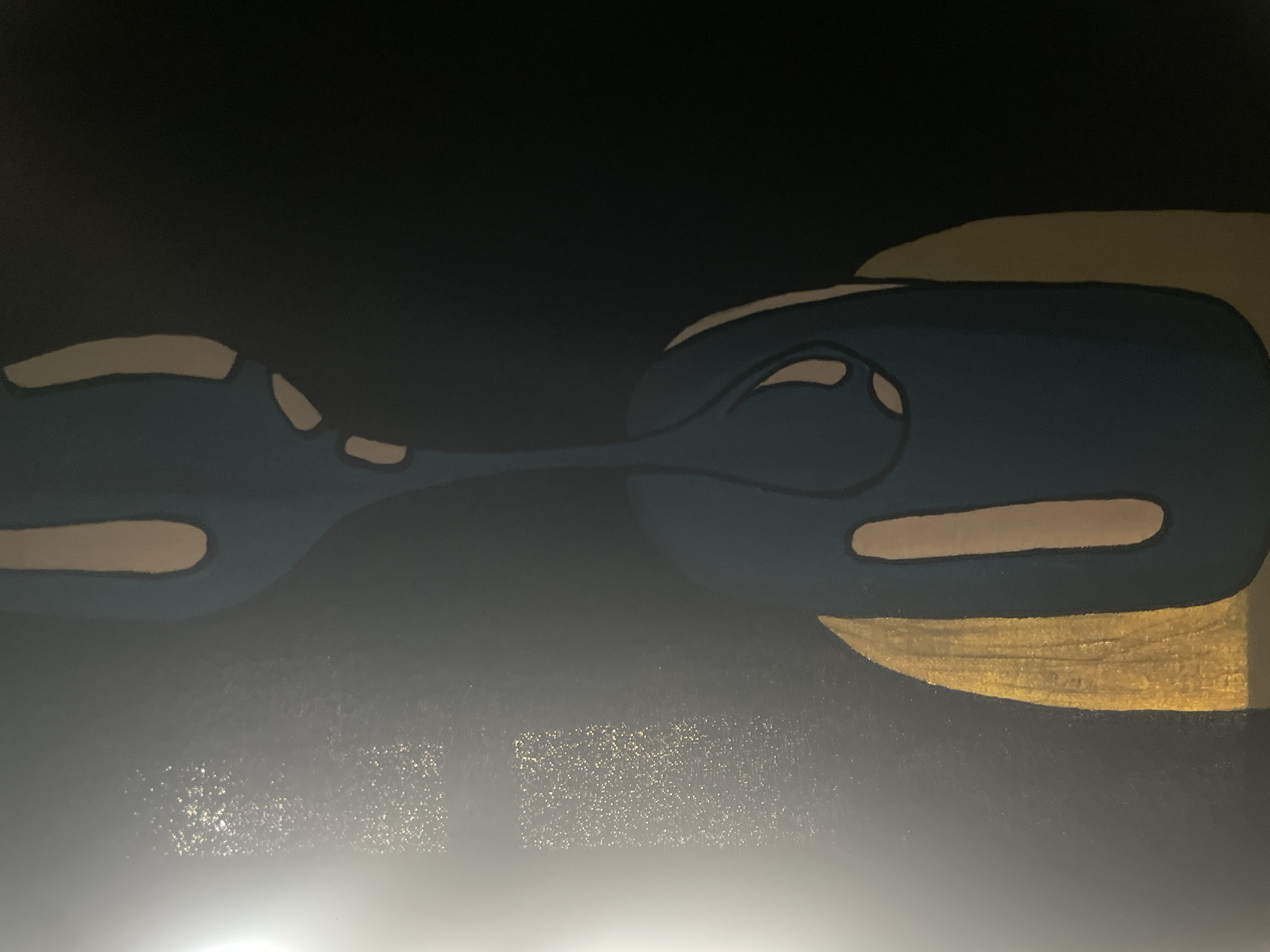 The image shows a close-up view of abstract artwork on a wall. The artwork features bold, stylized shapes in shades of blue, beige, and gold against a dark background. The dominant element is a large, dark blue, rounded shape with a slender, elongated extension reaching upwards, also in dark blue. Within these blue shapes are several irregularly shaped beige areas, creating a sense of depth and contrast. A prominent gold, sickle-shaped element is positioned to the lower left of the dark blue mass. The background is predominantly black, with a bright, diffused light source illuminating the left side of the image, creating a gradient effect and highlighting a textured area with small, reflective specks. The overall impression is that of a contemporary, minimalist painting, possibly with metallic accents. The provided location of Kurunduma, Nigeria, does not appear to be directly represented or depicted within the image itself; the image is solely focused on the artwork.