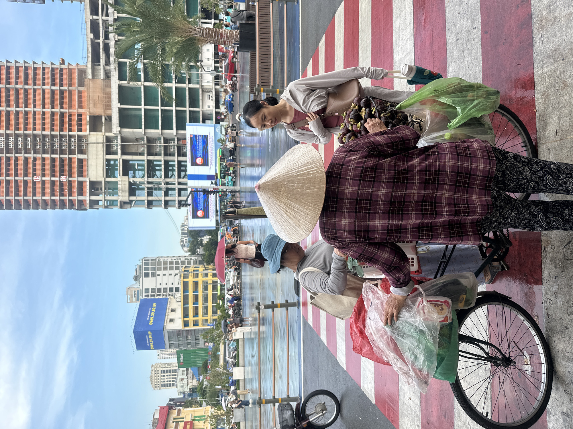In Thủ Đức City, Vietnam, on a wet street with a red and white striped pedestrian crossing, three individuals are gathered around a bicycle loaded with goods. A person wearing a traditional conical hat and a plaid shirt stands facing away from the camera, holding clear plastic bags filled with items. To their left, another person in a blue hat and grey long-sleeved shirt is also looking at the goods. To the right, a woman in a grey tracksuit and a brown shoulder bag is engaged in conversation with the person in the conical hat, looking at a basket of purple mangosteen fruits. In the background, unfinished high-rise buildings and a billboard with Vietnamese text are visible. The street is wet, indicating recent rain, and reflections of the surroundings can be seen on the ground. A motorcycle is parked to the left of the frame.