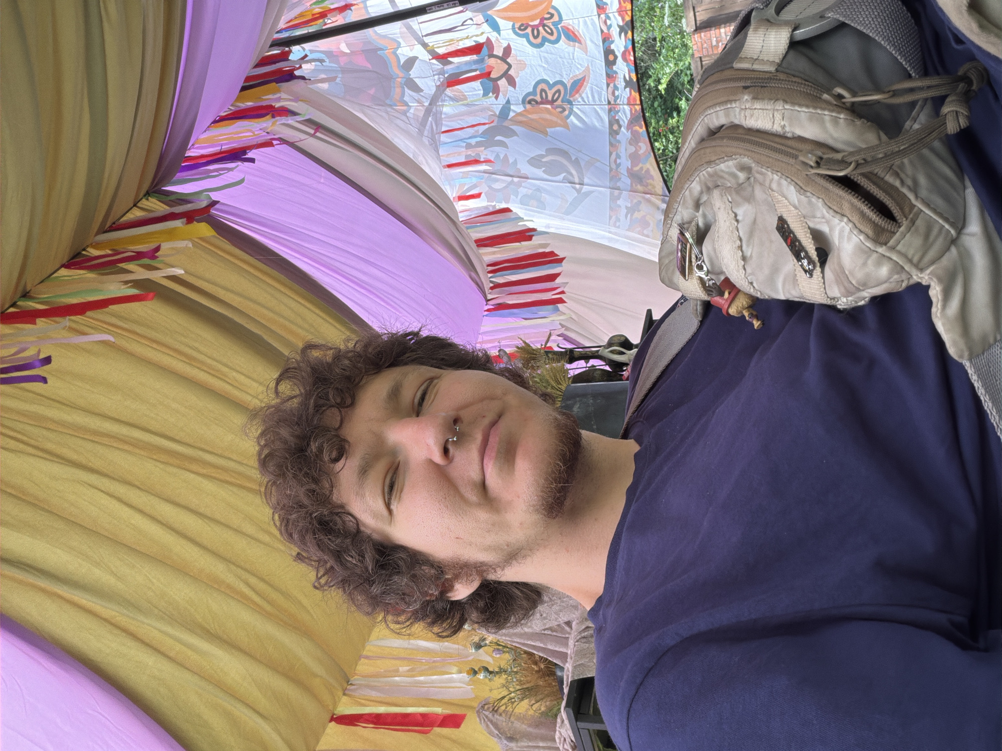 A young man with curly brown hair, a nose ring, and light stubble is pictured in a selfie, seated under a tent decorated with colorful ribbons and fabrics. The tent's canopy is a blend of yellow and purple hues, with various lengths of red, yellow, white, and purple ribbons hanging from the structure. The man wears a dark navy blue t-shirt and has a beige crossbody bag slung over his shoulder. The bag is adorned with several small charms, including what appears to be a red gnome-like figure. Behind him, a glimpse of foliage and a brick wall can be seen through an opening in the tent. The overall setting suggests an outdoor gathering or festival. The image location is identified as Shebanovo, Russia.