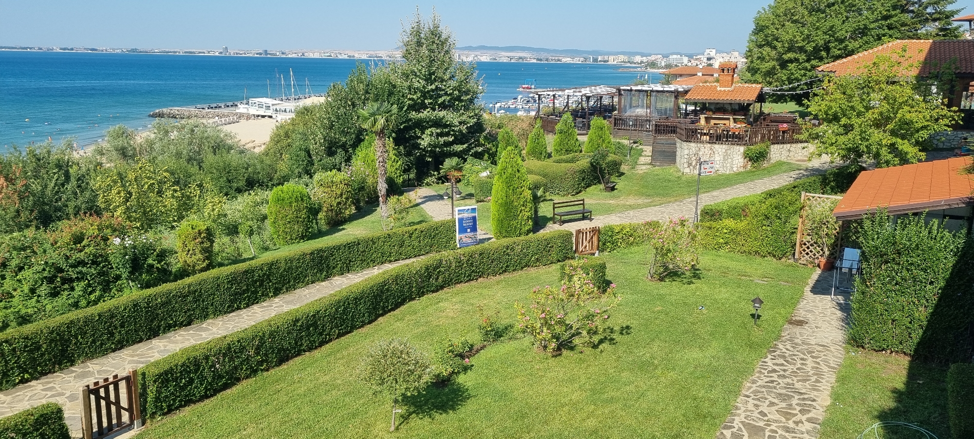 This image shows a scenic view of the coastline in Sveti Vlas, Bulgaria. The foreground features a meticulously manicured garden with lush green lawns, neatly trimmed hedges, and winding stone pathways. Several evergreen trees and bushes are strategically placed throughout the garden, adding texture and color. A wooden bench is visible along one of the paths, offering a place to rest and enjoy the surroundings. In the middle ground, a complex of wooden structures with red-tiled roofs appears to be a restaurant or resort area, with outdoor seating and umbrellas. Beyond this, the turquoise waters of the Black Sea stretch towards the horizon, dotted with sailboats and a pier. In the distance, the cityscape of Sveti Vlas is visible, characterized by a mix of modern buildings and traditional architecture. The sky is clear and blue, indicating a sunny day. There are no people visible in the frame.