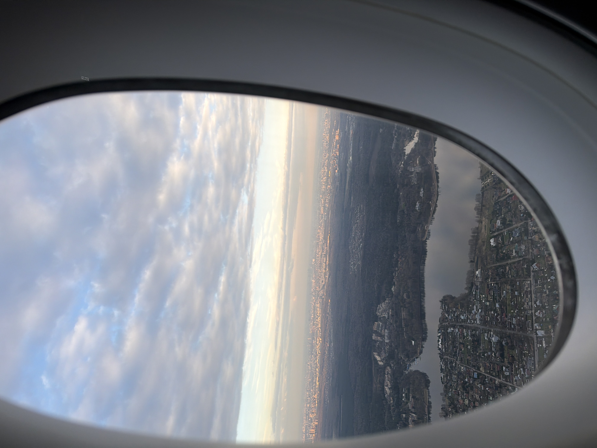 A view from an airplane window captures a landscape that includes clouds, a city skyline, a forested area, and a residential area. The clouds are visible on the left side of the image and appear to be light-colored. A city skyline is visible with closely packed buildings. A darker forested area stretches horizontally across the middle. A densely populated residential area with visible houses and streets occupies the right side of the window frame. The frame of the airplane window is visible, with a white inner lining.