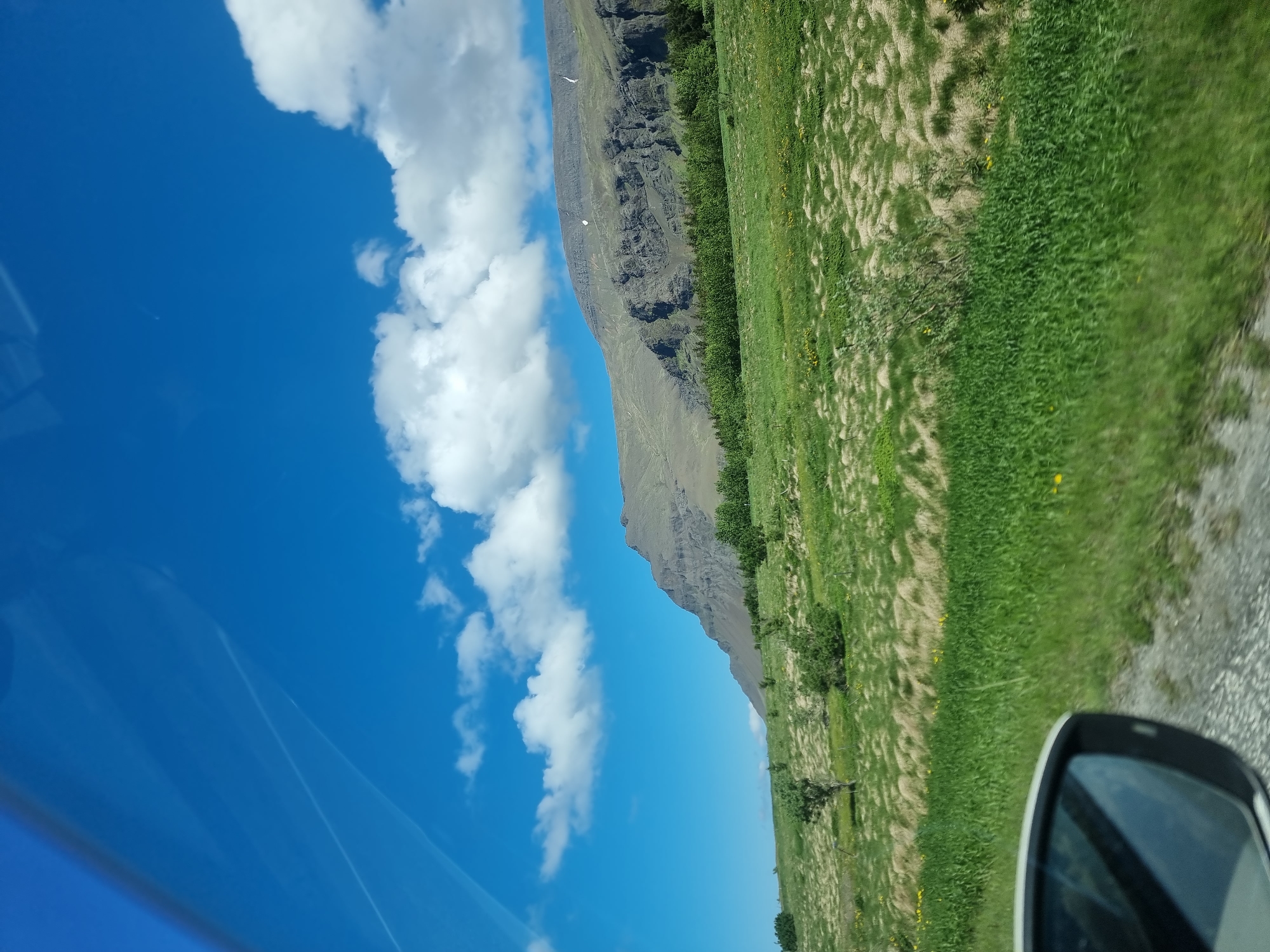 This image captures a scenic view from a car window, likely taken while driving along a rural road in Iceland, given the landscape. The foreground shows a portion of a side mirror of a car, reflecting the bright blue sky and a hint of the road ahead. The landscape features rolling green hills covered in lush grass and scattered patches of drier, lighter-colored vegetation. In the mid-ground, a line of dense green trees forms a distinct boundary. Behind the trees, a massive, layered mountain rises majestically, its slopes displaying intricate geological formations and rocky outcrops. The sky above is a vibrant blue, adorned with scattered white cumulus clouds, some of which cast soft shadows on the landscape below. There are no people or identifiable structures visible in the scene, emphasizing the natural and undeveloped beauty of the environment. The lighting suggests it is daytime, possibly late morning or early afternoon, with the sun casting clear light on the scene. The overall impression is one of vast open space and the raw, natural beauty of the Icelandic countryside. The location is unlikely to be within the city of Reykjavik itself, but rather in its surrounding natural areas, possibly on a route leading out of the capital.