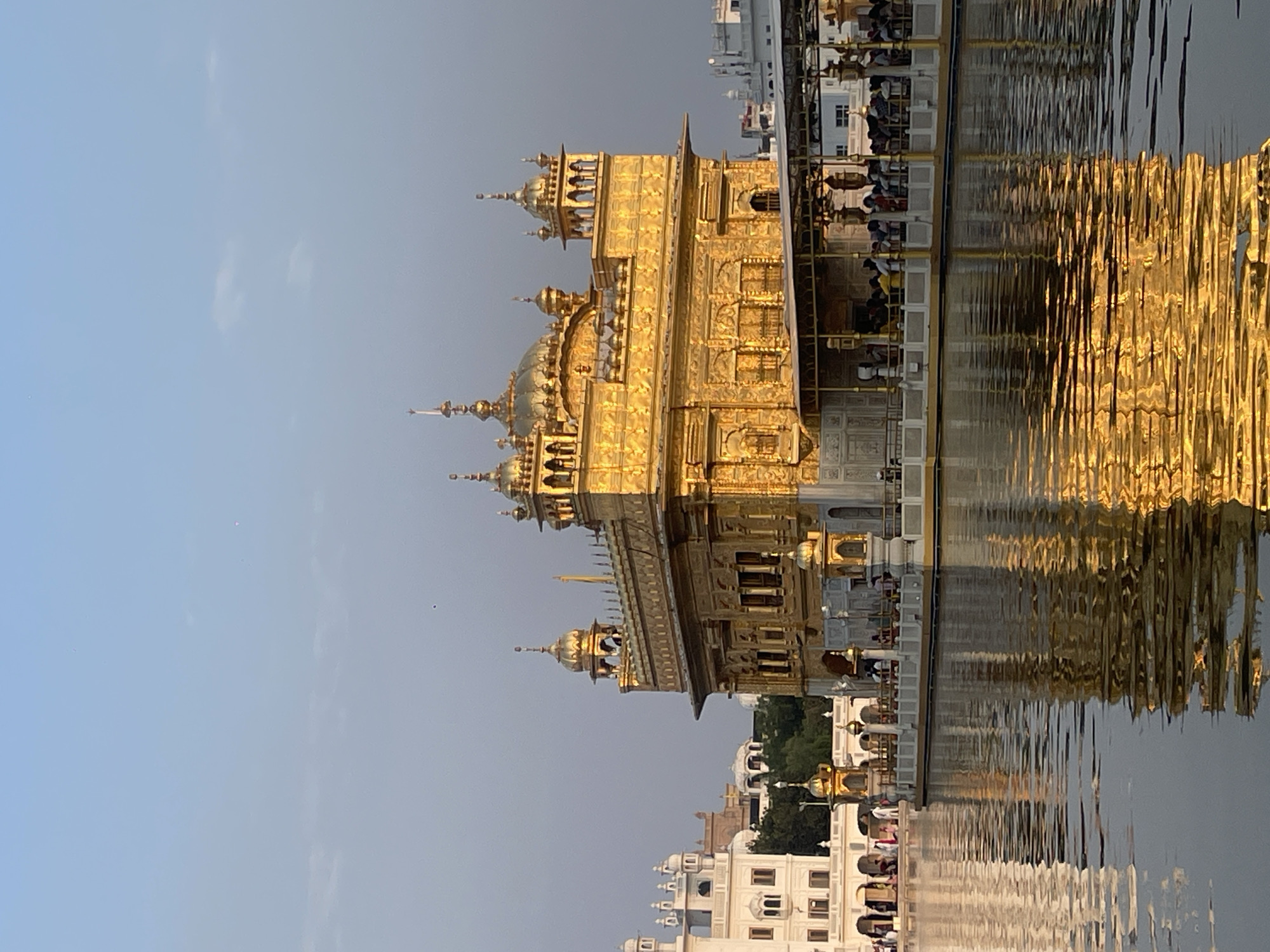 The image shows the Golden Temple, also known as Harmandir Sahib, in Amritsar, India. The temple, made of gold, is situated in the middle of a large body of water, reflecting its golden hues. The sky is clear with some faint clouds. Several people are visible on a walkway surrounding the temple and in the foreground, standing on a pier or walkway. The water surface has ripples, distorting the reflection of the temple. In the background, there are other buildings, some white and some with domes, and greenery. The overall scene is bathed in the warm light of what appears to be either sunrise or sunset, creating a serene and majestic atmosphere.