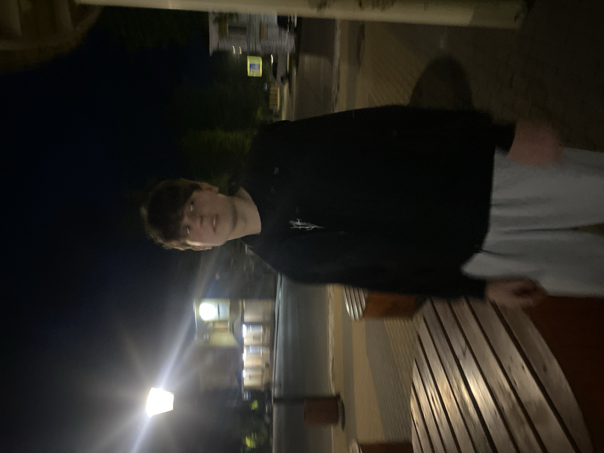 A nighttime, portrait-oriented photo shows a young male person, likely in their late teens or early twenties, looking towards the camera. The image is taken outdoors in a dimly lit environment.
The person occupies the left-center of the frame, from the chest up. They have dark, medium-length hair with bangs and are wearing a dark-colored hoodie or sweatshirt with a light-colored design on the front. Light-colored pants are visible below the waist. Their expression is neutral or slightly inquisitive.
To the right, a wooden, slatted surface, resembling a bench or a table, runs diagonally across the frame. Its surface is reflective, catching some of the ambient light.
The background is dark, indicating it is nighttime. Several artificial light sources illuminate parts of the scene. A prominent, bright streetlamp with a significant glare is visible in the lower-left, casting light upwards. Further in the background, dimly lit buildings can be seen, suggesting an urban or suburban setting. One building appears to have several windows lit up, and there's a faint indication of a street or pathway. The overall impression is that of a casual outdoor shot in the evening. No visible text is discernible in the image.