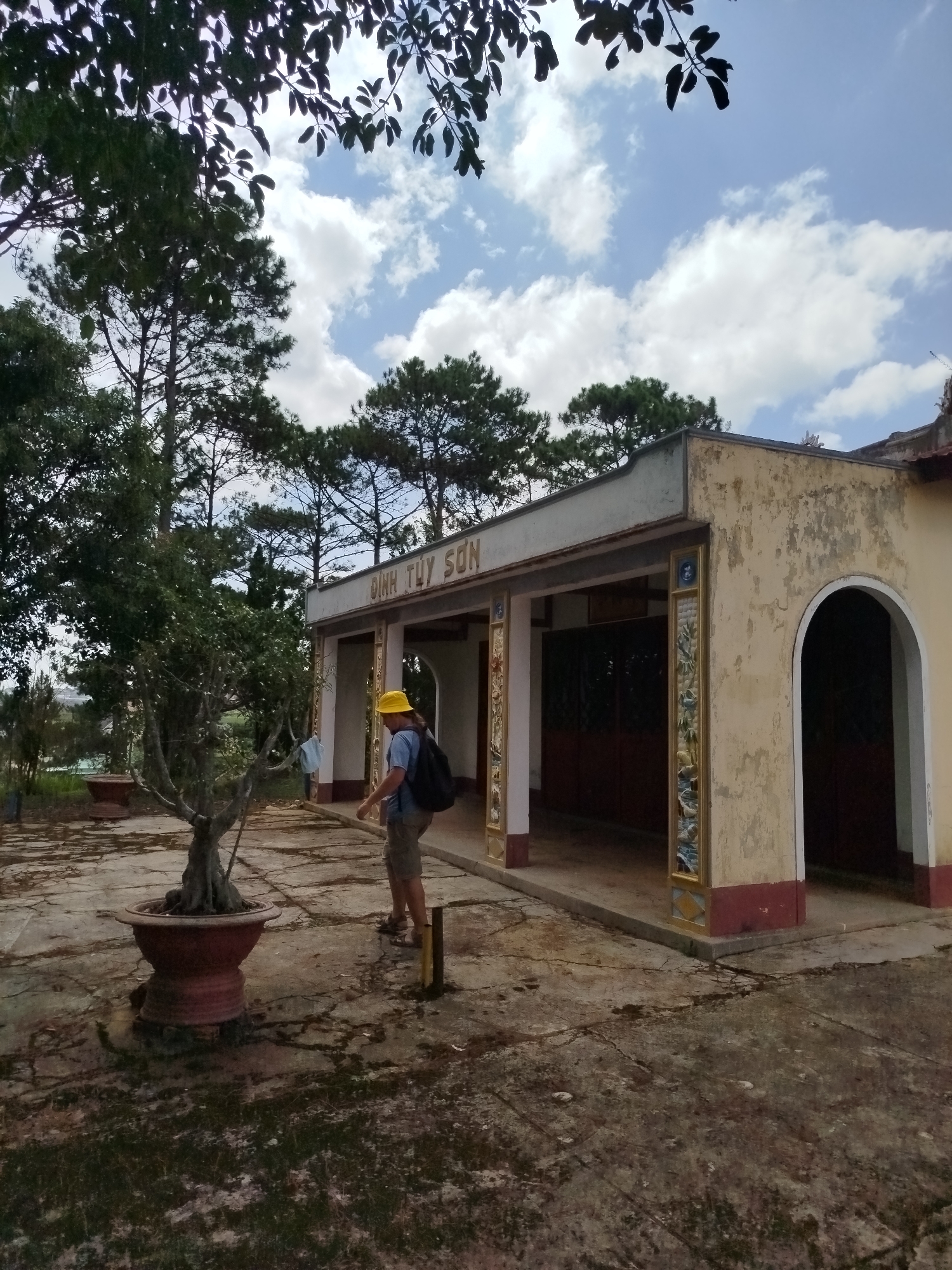 A person wearing a yellow hard hat and a blue shirt walks past a yellow building in Tuý Sơn, Vietnam. The building has a sign that reads "ĐÌNH TUÝ SƠN". The building appears to be a temple or a historical site, with decorative pillars and an arched entrance. The ground in front of the building is paved with cracked concrete. A potted bonsai tree sits to the left of the building, and various trees and foliage surround the scene. The sky is blue with scattered clouds. The person is carrying a black backpack and appears to be walking away from the camera.