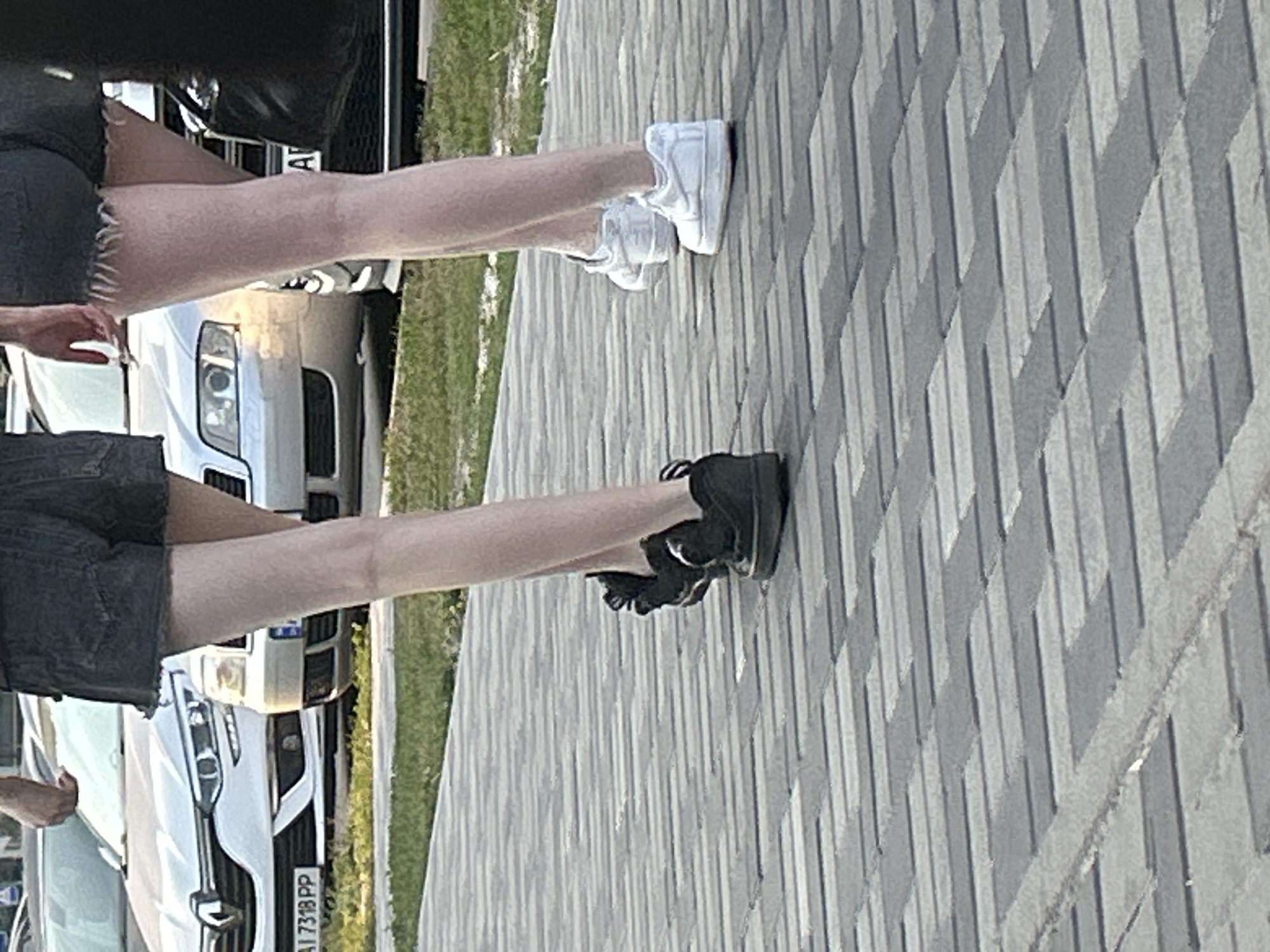 Two individuals, seen from the waist down, are walking on a patterned sidewalk in Brovary, Ukraine. The person on the left wears black shorts and black sneakers. The person on the right wears dark denim shorts with frayed edges and white sneakers. Behind them, a white Renault car with a license plate reading "AI 7318 PP" is parked, along with a silver Audi. The environment appears to be an outdoor urban setting with green grass and a paved walkway.