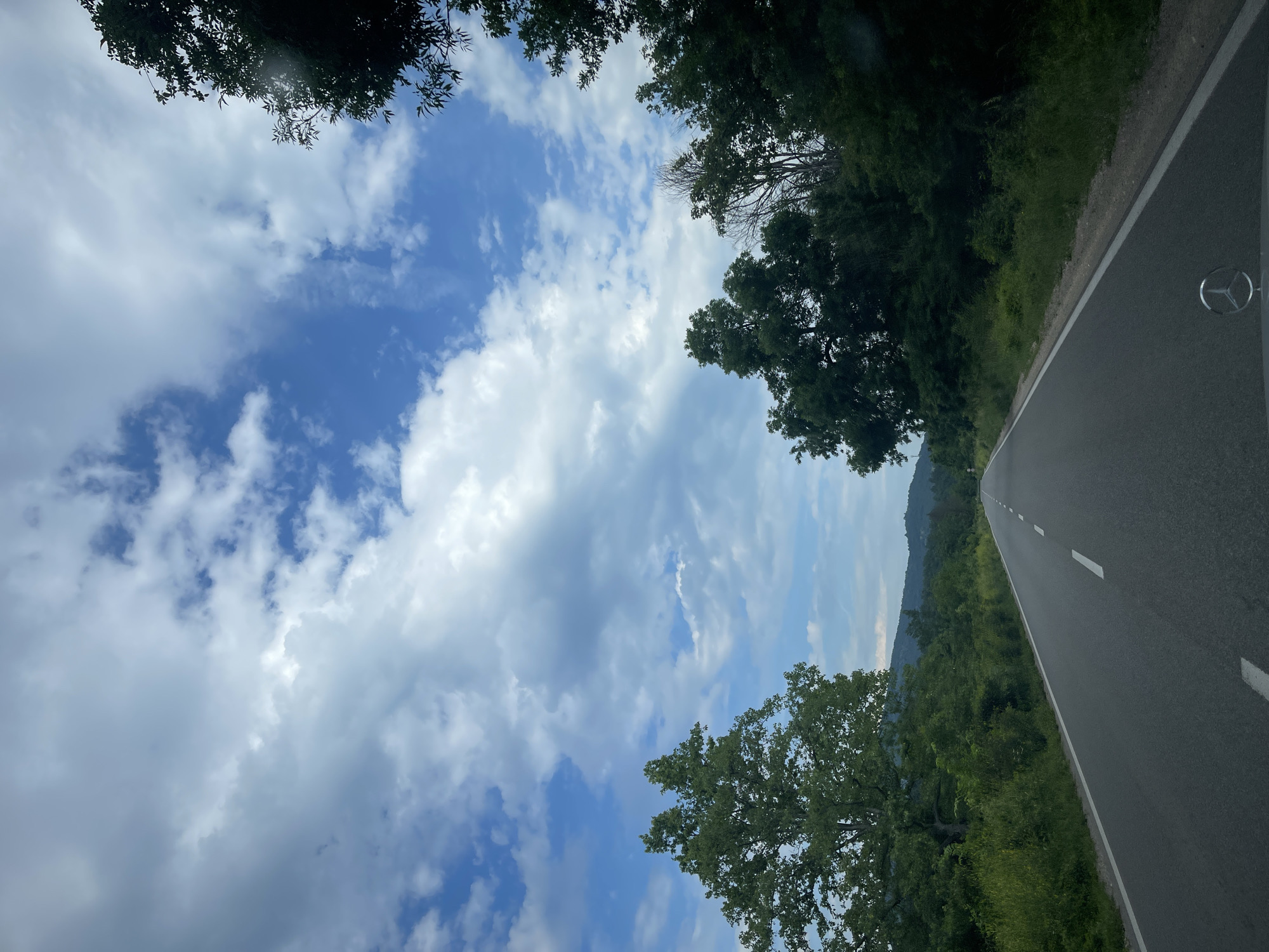 The image depicts a two-lane asphalt road stretching into the distance, flanked by dense green trees and vegetation. The sky is a mix of blue and white clouds. The scene appears to be a rural setting in Komarevo, Bulgaria, as suggested by the presence of rolling hills in the background. A portion of a car's hood with a Mercedes-Benz logo is visible in the foreground. There are no people or animals visible in the image. The overall impression is one of a quiet, scenic drive through the countryside.