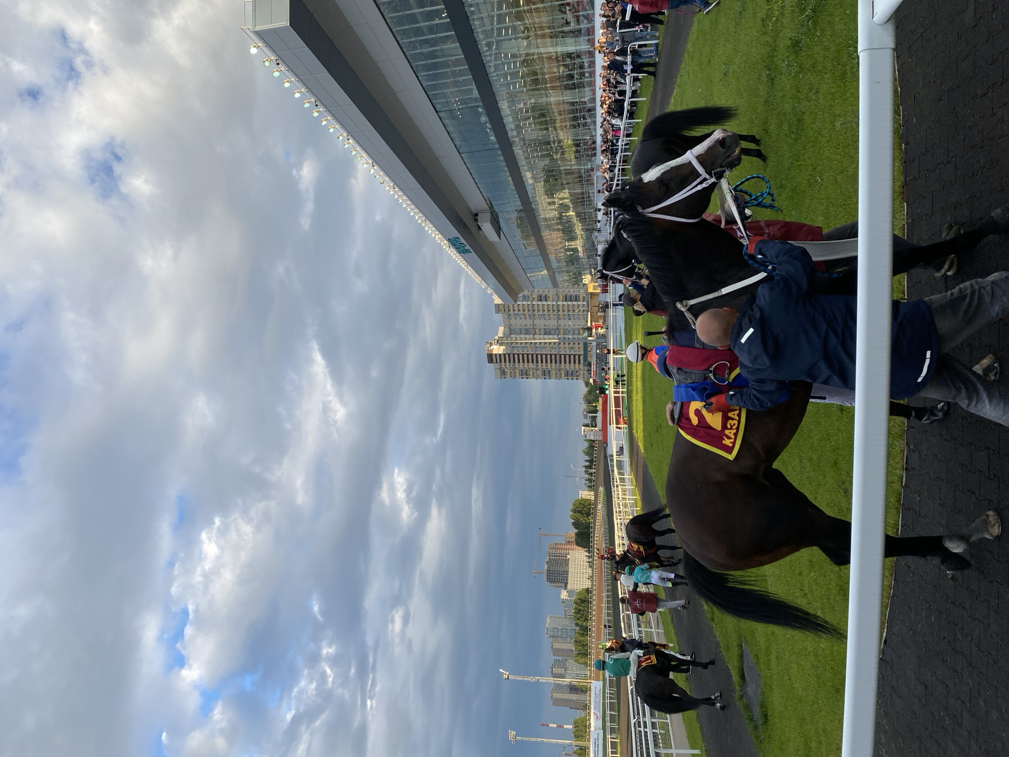 The image depicts a horse race track in Kazan, Russia, with a large, modern stadium in the background. Several horses and jockeys are visible on the track, with trainers or handlers tending to them. The sky is cloudy, with patches of blue sky peeking through. The stadium has a sleek, contemporary design with extensive glass facades and numerous lights along its roofline. In the distance, city buildings and construction cranes can be seen, indicating an urban environment. The scene suggests a sporting event in progress or preparing to commence.