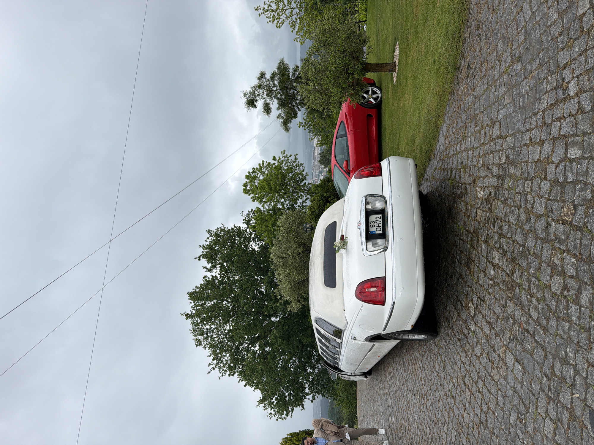 The image captures a scene in Vila Real, Portugal, likely in a semi-rural or residential area. A white limousine is parked on a cobbled stone surface, possibly a driveway or a side street. Behind the limousine, a red car is partially visible on a patch of green grass next to a tree. The sky is overcast, indicating potentially cloudy or rainy weather. Power lines are visible crossing the upper part of the frame. In the lower part of the image, two people are standing and possibly interacting, though the details are limited due to the angle and distance. The environment includes trees, suggesting a residential or semi-rural setting within Vila Real. The focus is primarily on the vehicles, with the limousine appearing to be a central subject of the image.