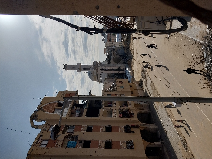 A partially destroyed building is visible on the left, with visible damage and exposed interiors. A mosque with a tall minaret stands in the distance. A road runs between the building and the mosque, with several people walking. Debris and rubble are scattered along the roadside. Overhead wires and poles are present, casting shadows on the ground. The sky is partly cloudy.
