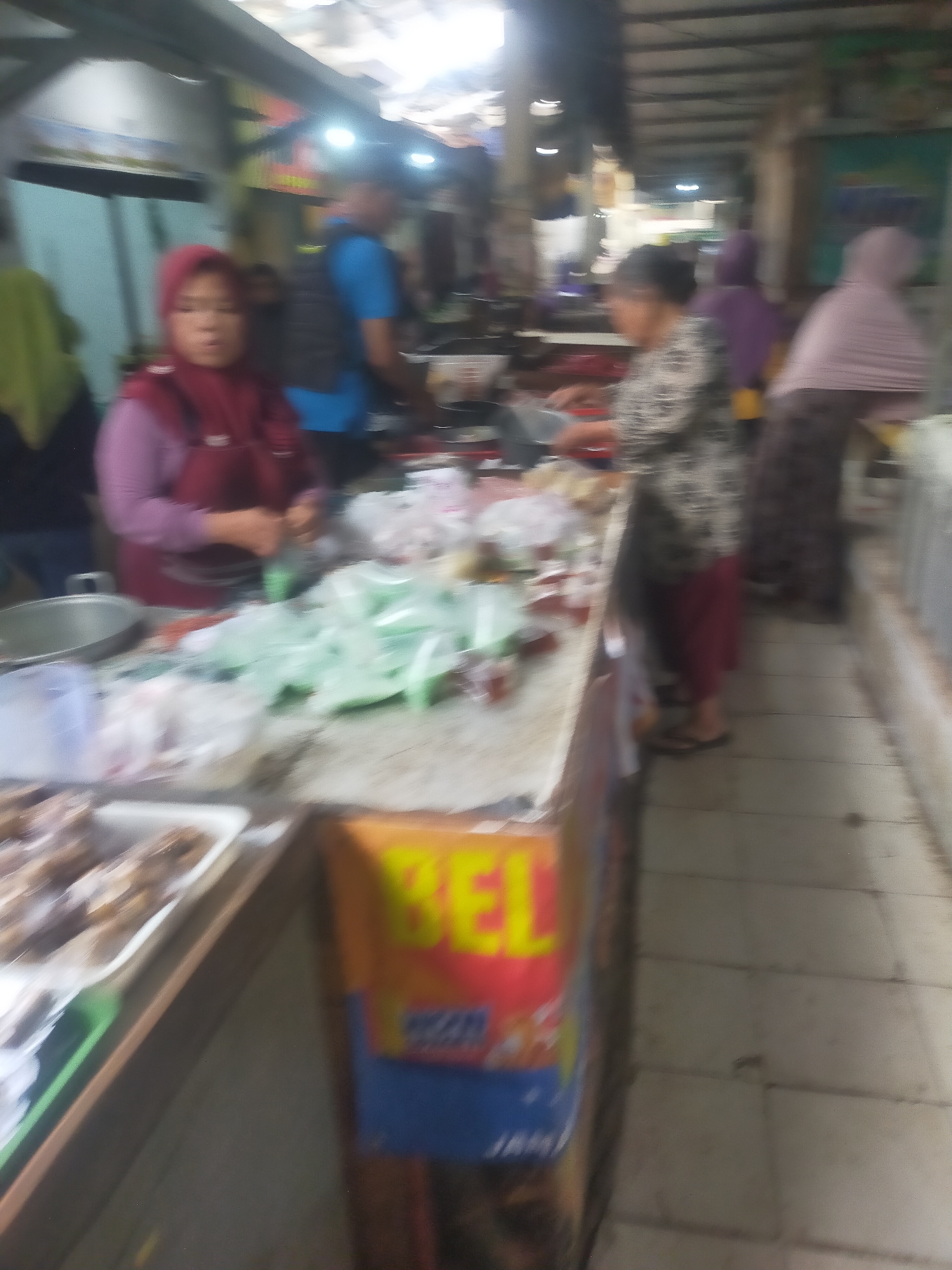 The image depicts an indoor market scene, likely in Trayeman, Indonesia. In the foreground, a woman wearing a red headscarf and a dark red apron is standing behind a counter, appearing to be handling some items. To her right, there are several plastic bags filled with green products. Further down the counter, another woman in a patterned shirt and maroon pants is visible, and beyond her, another individual wearing a light purple headscarf is seen. The market environment appears to be covered with a corrugated metal roof. The floor is tiled. Signs with Indonesian text are present on the counter and in the background. The lighting is somewhat dim, with some sources of light visible from above.