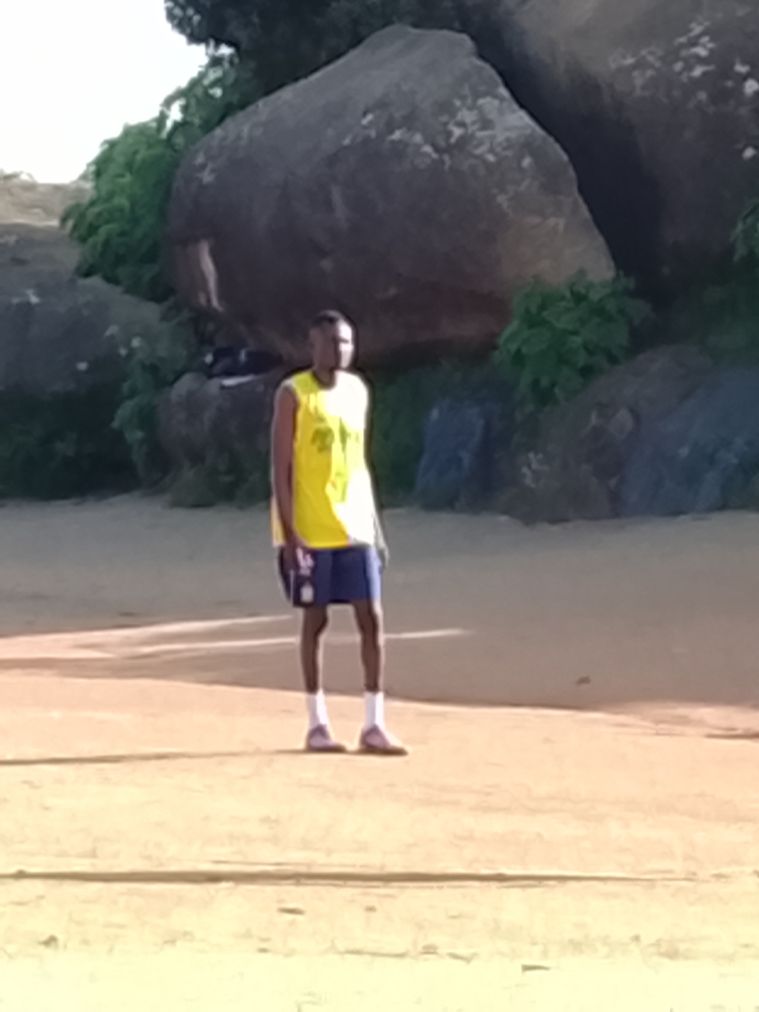 A medium shot captures a young Black male standing outdoors in a dirt field. He is wearing a bright yellow sleeveless jersey over dark blue shorts and white socks with pink athletic shoes. The man is facing slightly to the right of the camera, with his head turned to look directly at the viewer. His expression is neutral.
Behind him, large, rough-textured grey boulders are visible, partially obscured by dense green foliage. The ground is composed of dry, light-brown earth with some visible shadows. The lighting suggests it is daytime.
While the image does not provide specific landmarks within Jos, Nigeria, the presence of boulders and vegetation is consistent with the natural landscape found in and around the city, which features rocky outcrops and areas of greenery. The scene appears to be an open, natural space, potentially a park or a less developed area, within the city of Jos.