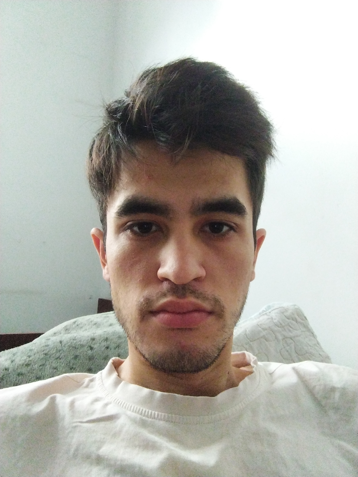 A young man with dark, tousled hair and a light beard is looking directly at the camera. He is wearing a plain white t-shirt. The background is a plain, light-colored wall, with a glimpse of a patterned pillow or blanket behind him. The lighting is bright and appears to be natural, casting a soft glow on his face. The overall setting suggests an indoor environment, possibly a home. The image is a close-up portrait, focusing on the man's face and upper torso. There is no indication of the city of Tashkent, Uzbekistan, within the image itself.