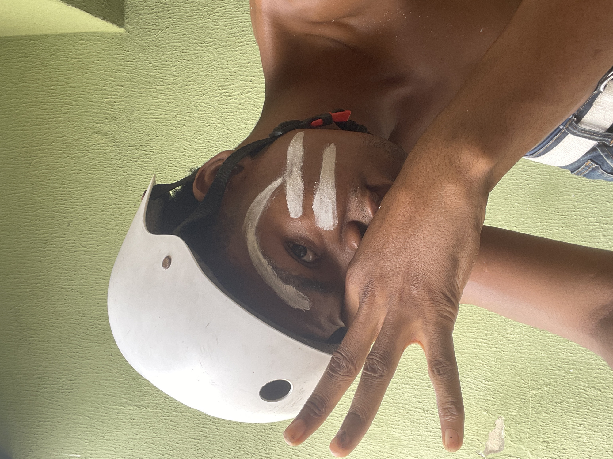 The image depicts a person with dark skin, wearing a white helmet. White markings are visible on the face. The person's hand is raised, with fingers spread. The background appears to be a textured green wall. The location is presumed to be Ado, Nigeria.