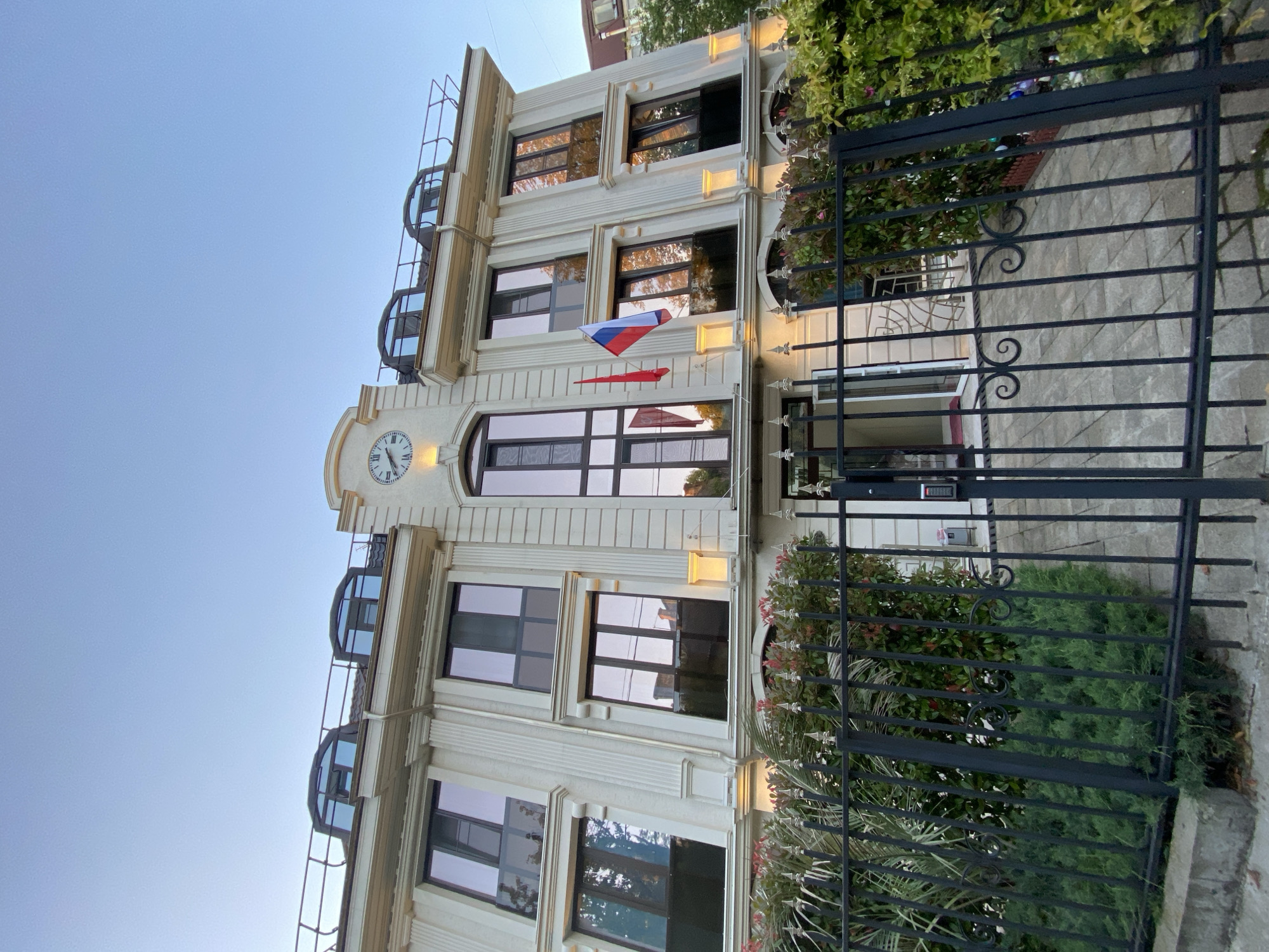 The image captures a stately, multi-story building under a clear, light blue sky, suggesting daytime. The setting is framed by a black wrought-iron fence in the foreground, adorned with decorative curled elements and enclosing lush green bushes and plants. An intercom or keypad is visible on a section of the gate.
The building itself features a light-colored, possibly beige or cream, facade with classical architectural details like pilasters and cornices. Numerous large rectangular windows with dark frames reflect parts of the sky and surrounding greenery. On the central axis of the building, above the main entrance area, a prominent clock face with Roman numerals is integrated into the architecture. The clock's hands indicate approximately 10:10.
Just below the clock, two flags are vertically displayed. The upper flag is clearly identifiable as the Russian national flag with its white, blue, and red horizontal stripes. The lower flag is predominantly red. Wall-mounted light fixtures are interspersed on the facade between floors, and the upper levels feature distinctive semi-circular window or balcony structures with metal railings. Given the location context of Sochi, Russia, and the display of the Russian flag, this building likely serves an official or diplomatic purpose. No people or specific activities are visible, presenting a calm and static scene.