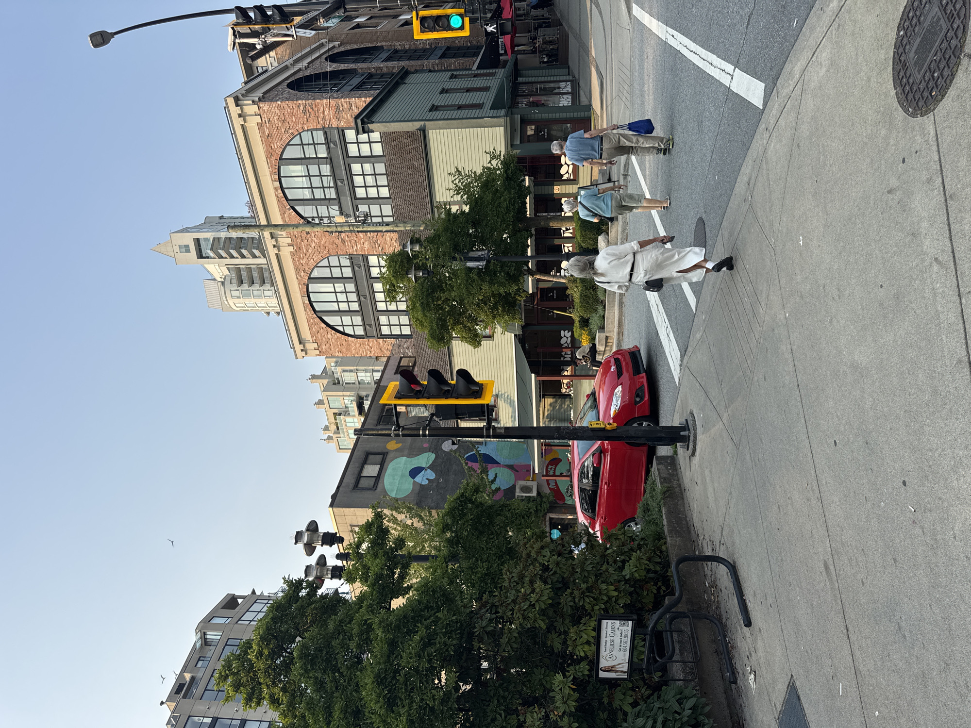 The image depicts a street scene in North Vancouver, Canada, likely in a downtown area characterized by a mix of older and modern architecture. In the foreground, a sidewalk runs alongside a street where a red hatchback car is parked. A group of three pedestrians, two women and one man, are walking away from the viewer on the sidewalk. The woman in front is wearing a white dress, the middle person a blue top and shorts, and the man a blue shirt and khaki pants. They are all facing away from the camera, walking across a crosswalk.
The street is lined with buildings of varying architectural styles. To the left, a building with a large green tree in front of it displays a colorful mural. Further down the street, an older brick building with large arched windows is visible, as is a more modern building with multiple stories. Traffic lights are present at the intersection, with one showing a green light. Street lamps are also visible. The sky is clear and blue, suggesting a daytime scene with fair weather. The overall impression is of a calm, urban environment.